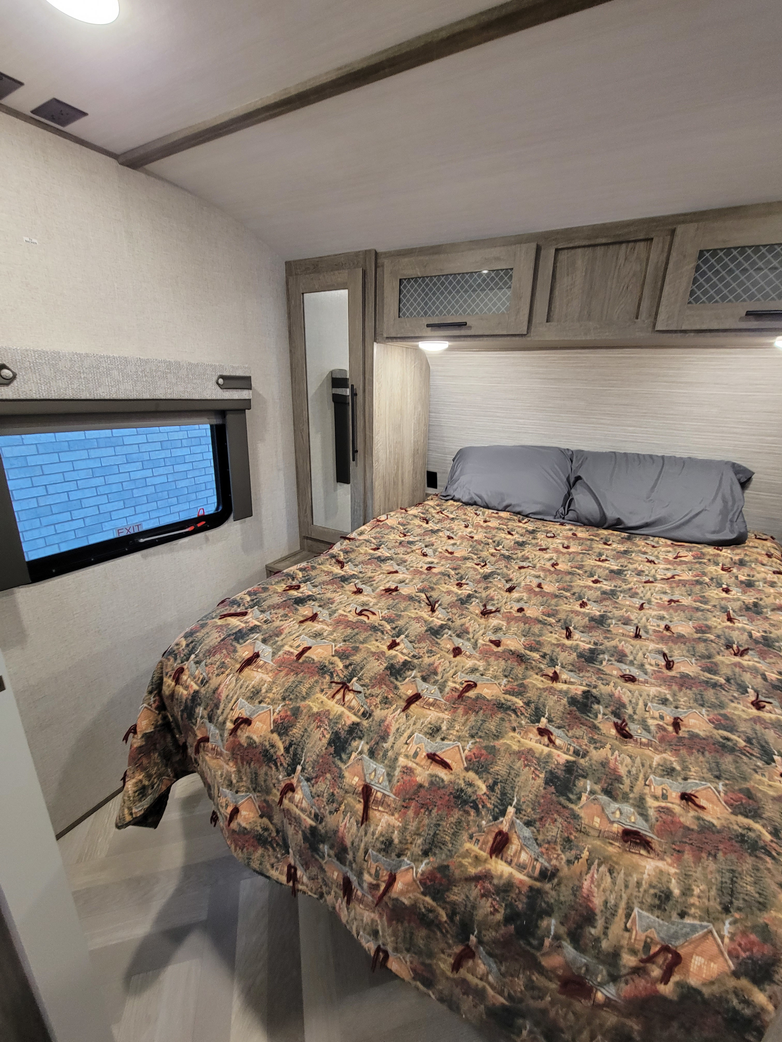 Cozy RV rental bedroom features a bed with a house and tree patterned comforter, gray pillows, overhead cabinets, and a mirrored closet.. Forest River Wildwood Heritage Glen 2021