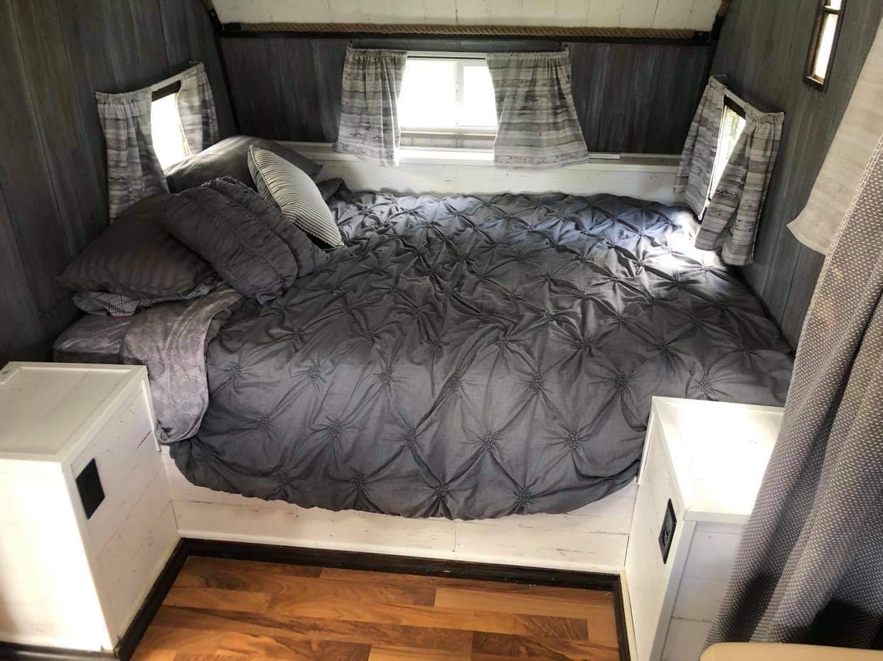 An inviting RV rental bedroom. Features a spacious gray tufted bed, multiple pillows, and side windows with curtains.. Nomad Rear fold 1962