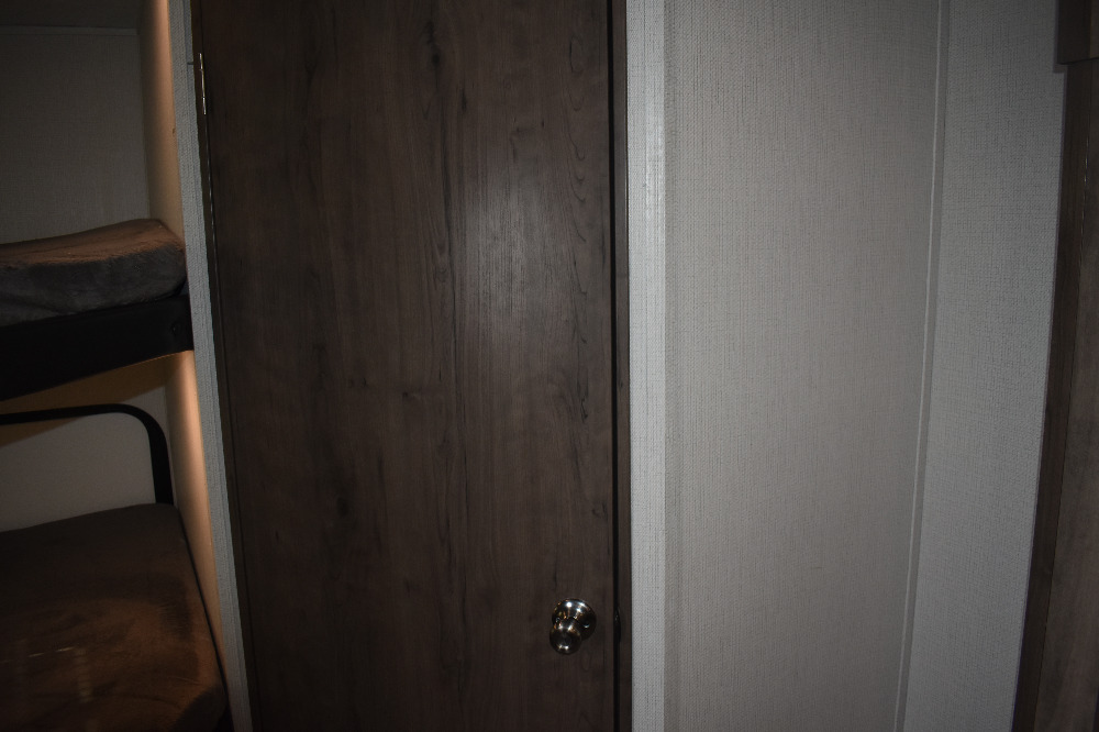 Inside an RV rental, showing a dark wood door with metallic knob, textured light walls, and bunk beds with a soft blanket.. Keystone Passport 2021