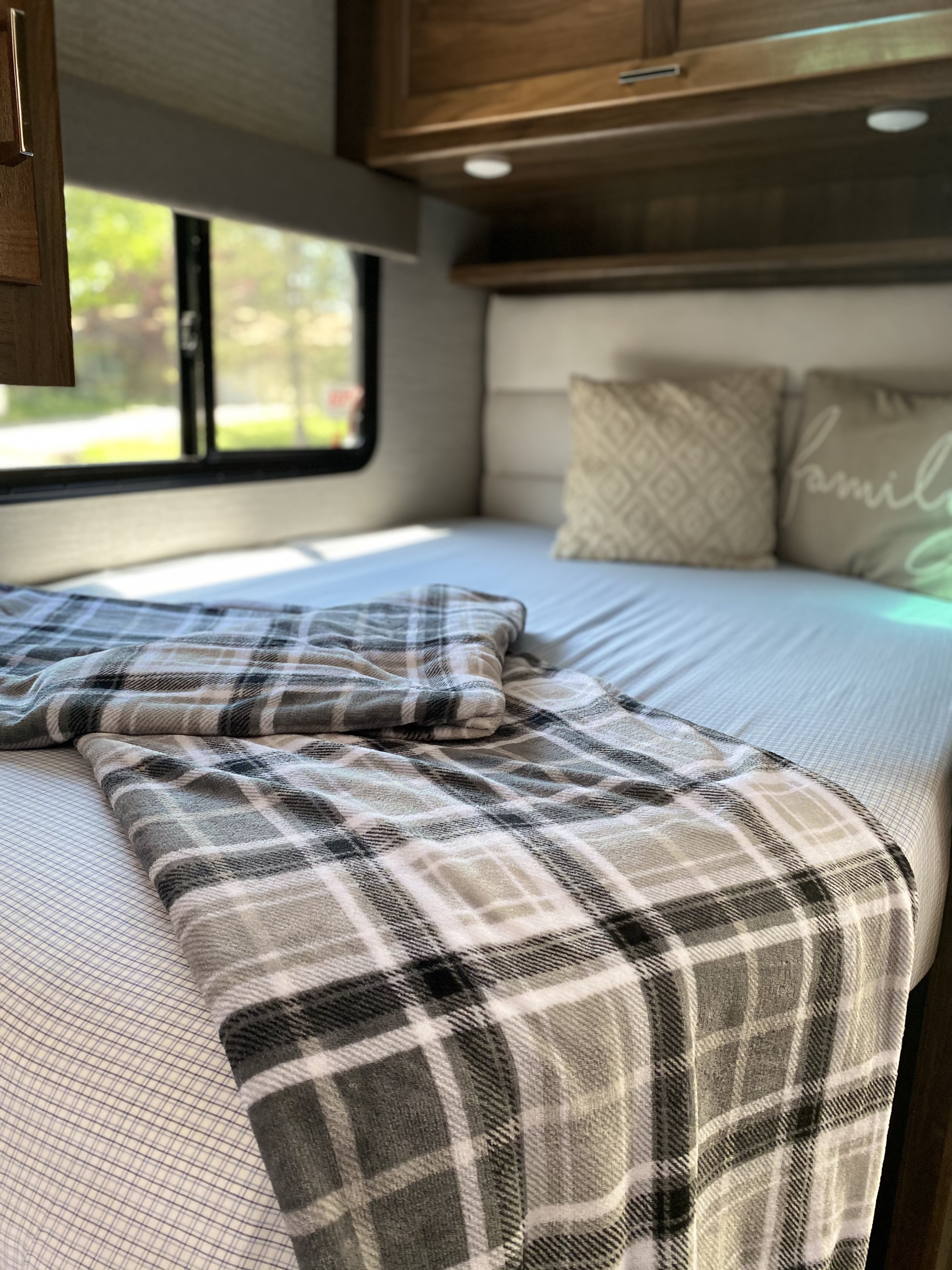 Cozy RV rental bed with plaid blanket, decorative pillows, & window view of nature outside.. Winnebago Minnie Winnie 2020