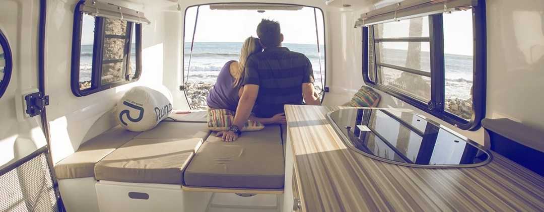 Inside an RV rental, a couple relaxes, admiring the ocean view from the open back. Ideal for beach getaways and adventures.. Happier Camper Happier Camper 2016