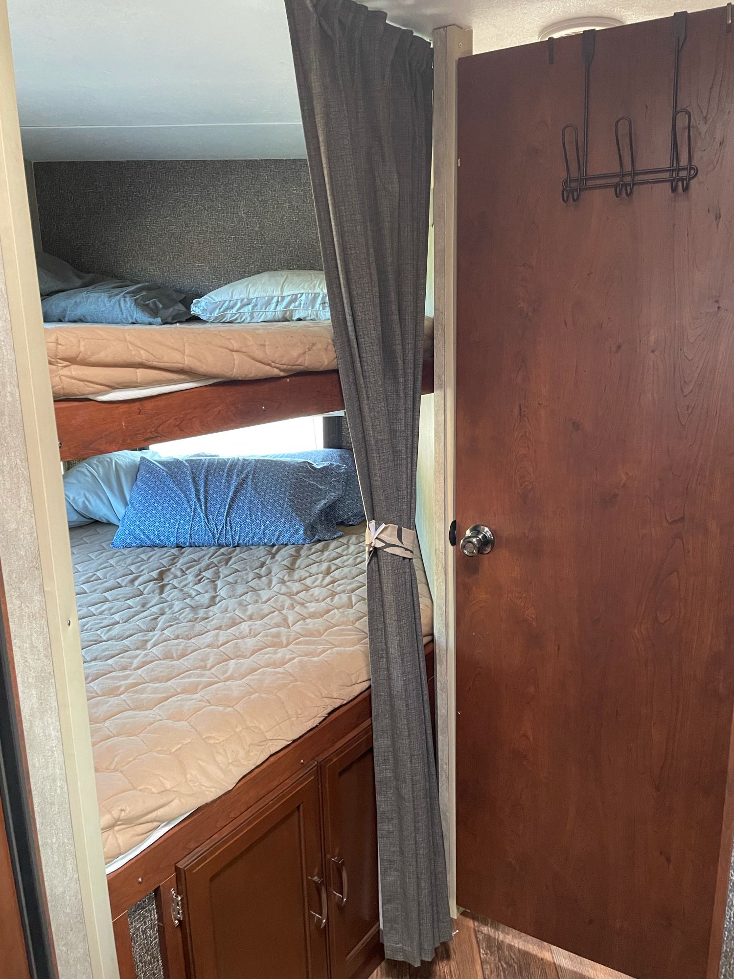View of an RV rental's cozy interior, showing three bunk beds with bedding and a patterned pillow, next to a wooden door.. Keystone Passport 2016