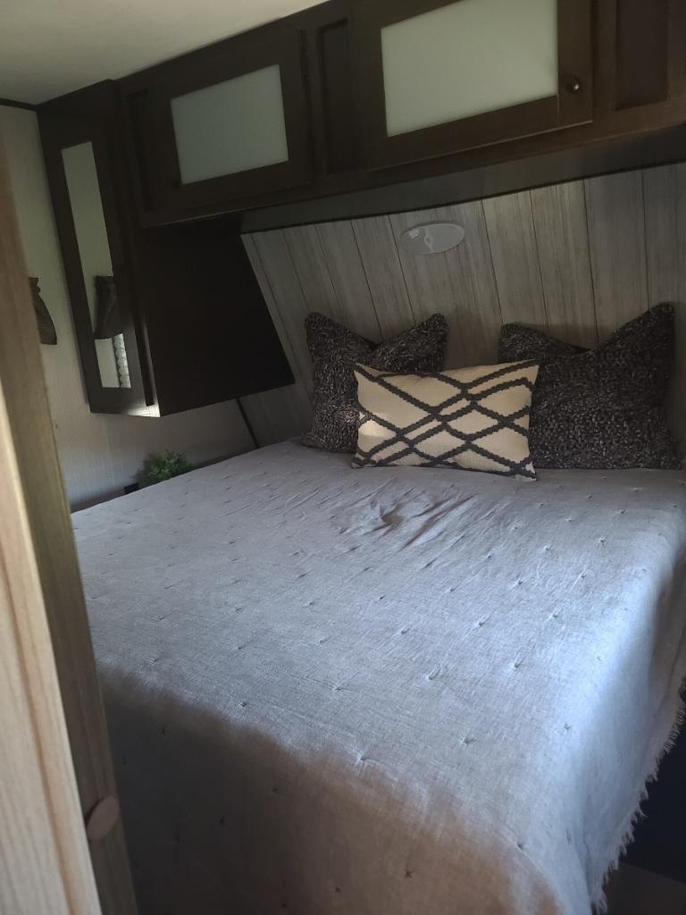 RV rental bedroom: cozy bed with grey quilt, pillows, wooden headboard, and dark overhead cabinets.. Heartland Pioneer 2021