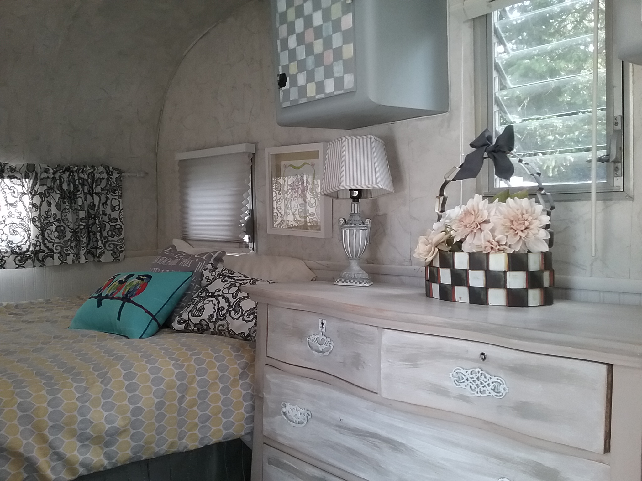 Inside a charming RV rental: a comfy bed with patterned bedding, a distressed dresser holding a lamp and floral decor.. Vintage Trailers Aloha 1959