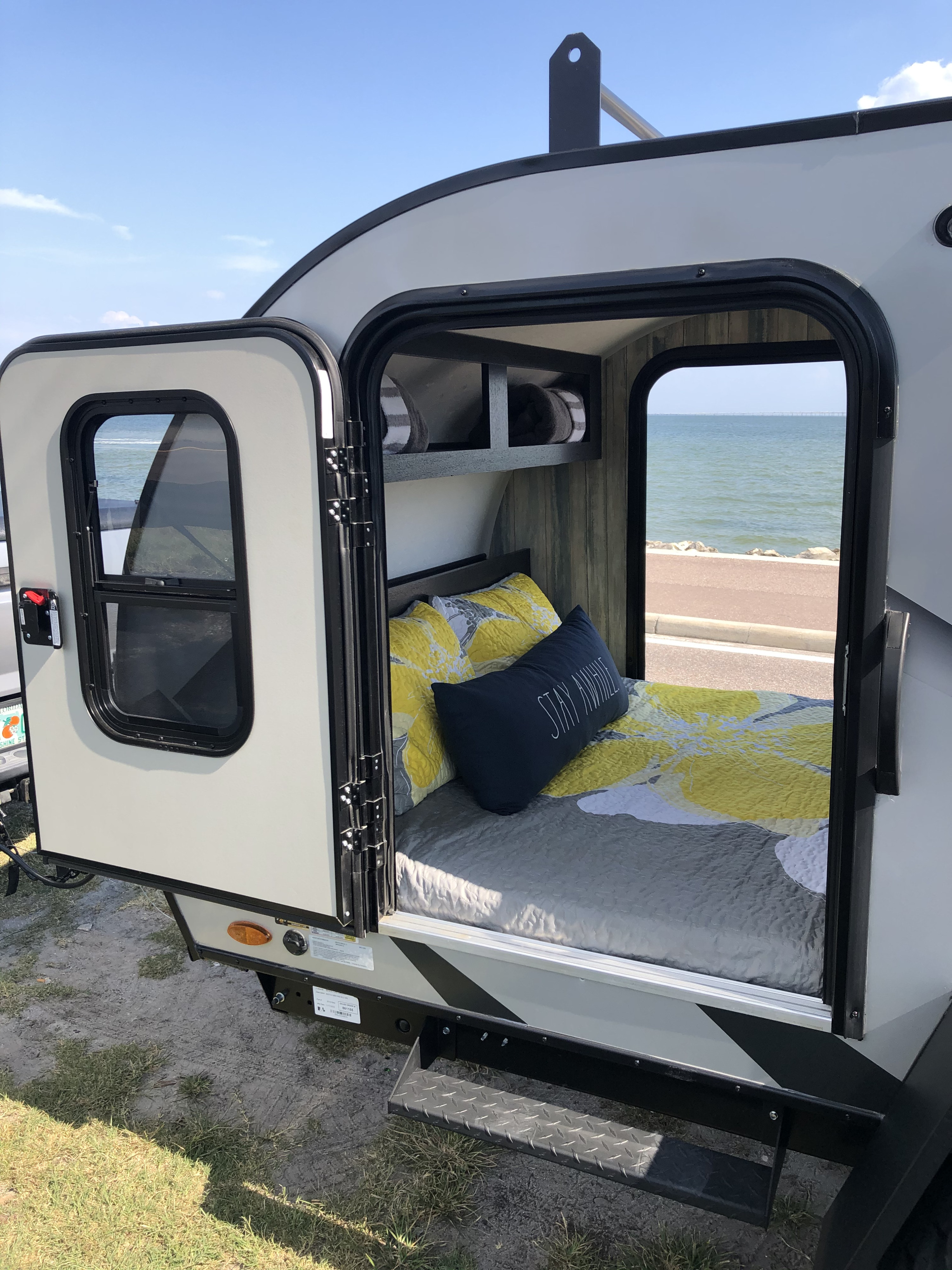Cozy rv rental teardrop camper bedroom with 'Stay Awhile' pillow, open to ocean views and coastal road.. Braxton Creek Free Solo 2021