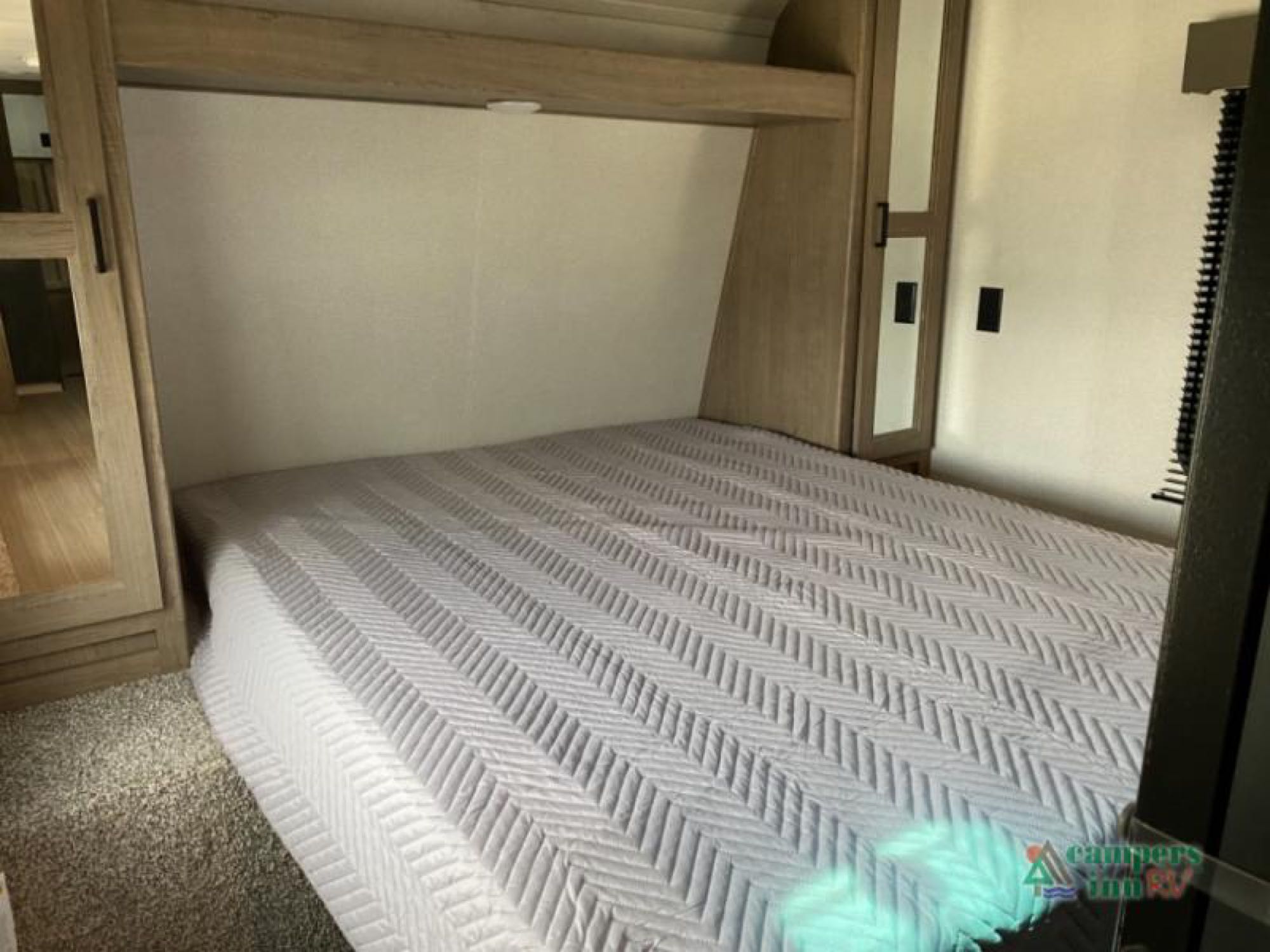 Interior of an RV rental bedroom showing a bed with a light gray quilted blanket, overhead shelf, and wooden cabinets.. Keystone Hideout 2021