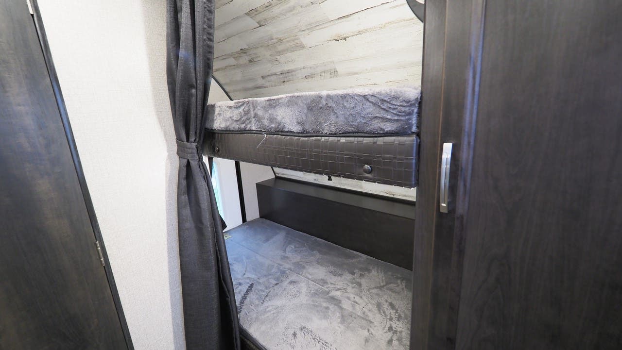Inside an RV rental, showing bunk beds with grey mattresses, a tied curtain, and dark wood cabinetry for efficient living.. Jayco Jay Feather 2021