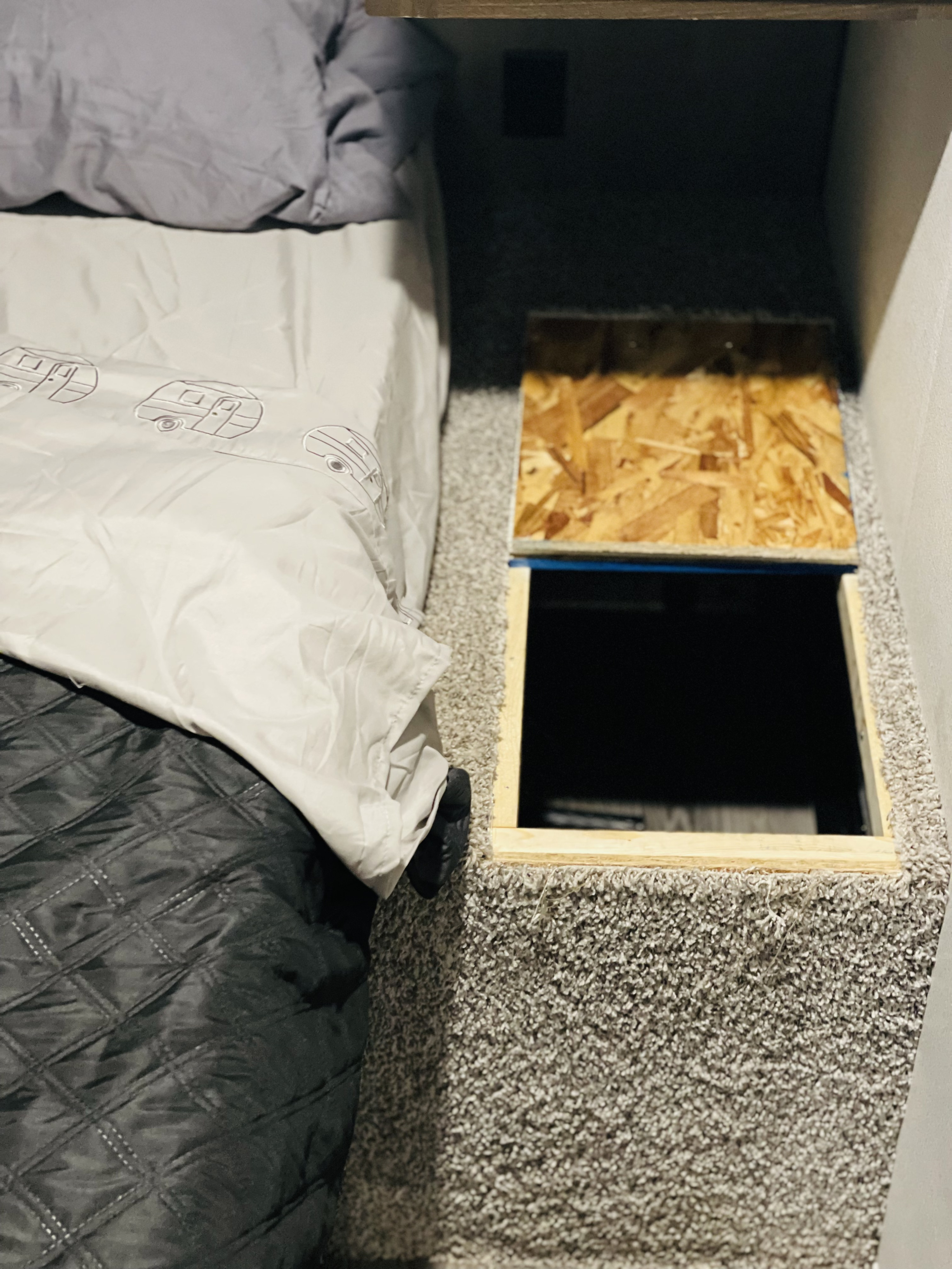 RV rental interior: Bed with camper duvet, adjacent open underfloor storage with a wood lid in the carpeted floor.. Keystone Springdale 2021