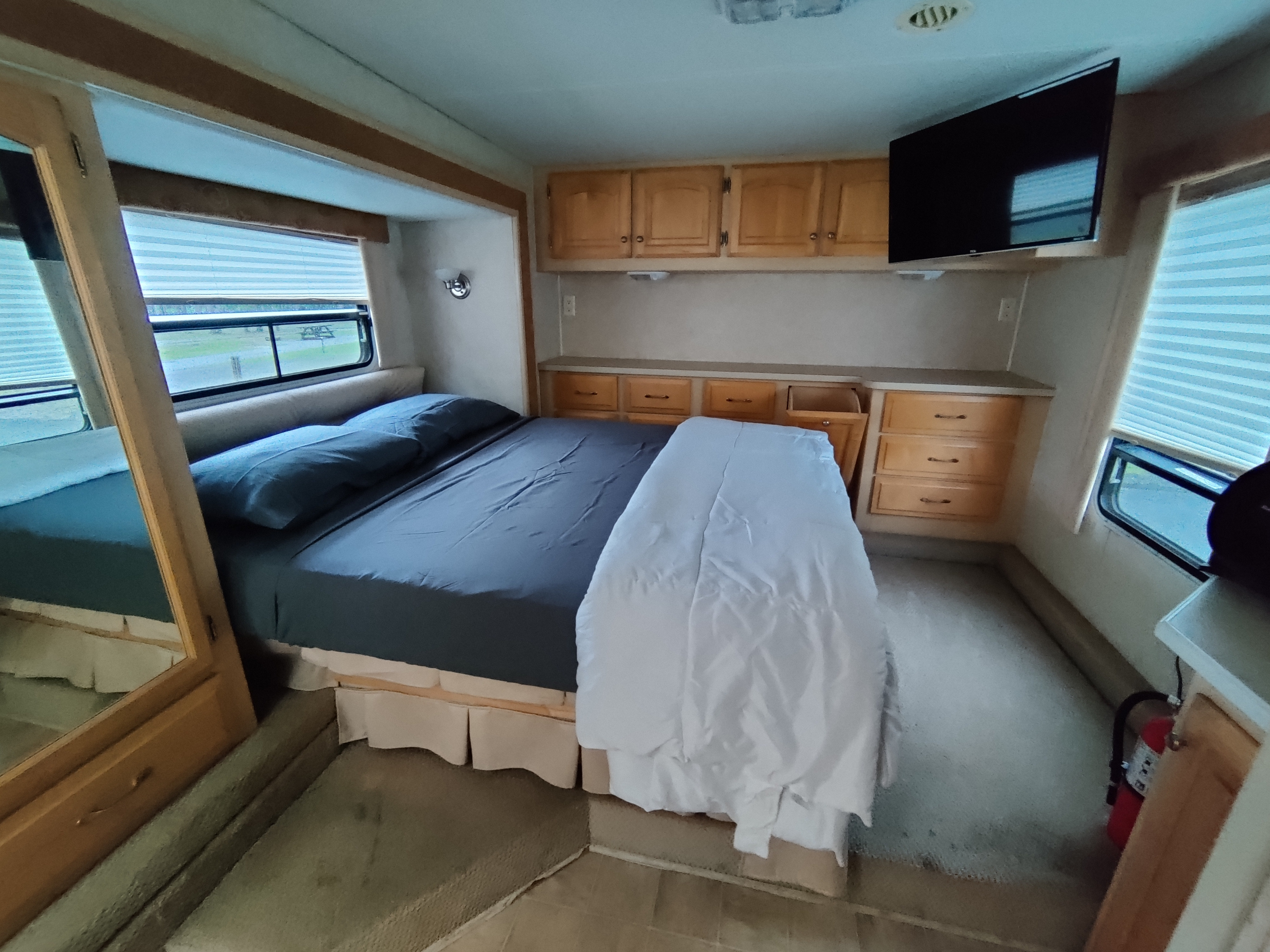 Relax in this RV rental bedroom: full bed, wood cabinets, wall-mounted TV, windows with blinds, and a fire extinguisher.. Carriage Cameo 2004