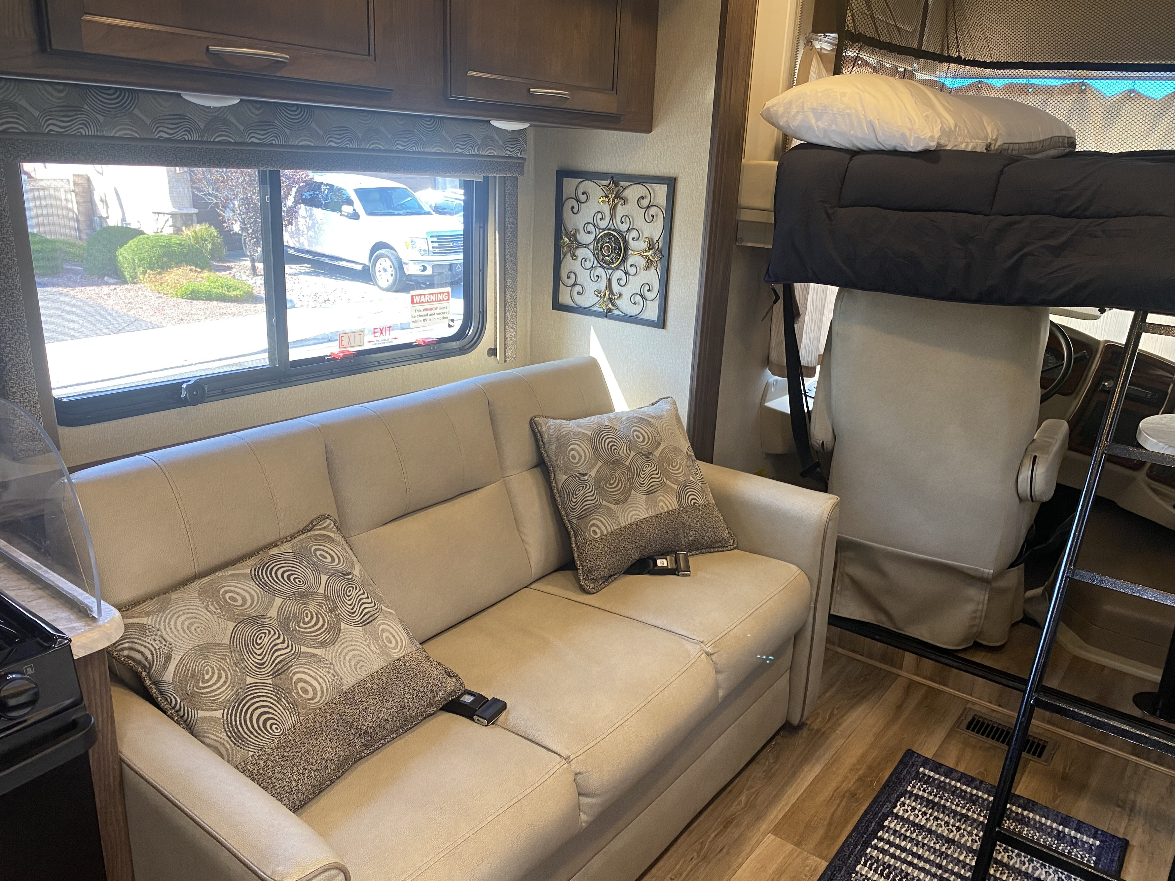 Inside an RV rental, showcasing a tan sofa with patterned pillows, an overhead bunk bed, and scenic window views.. Forest River Coachmen Pursuit 2019