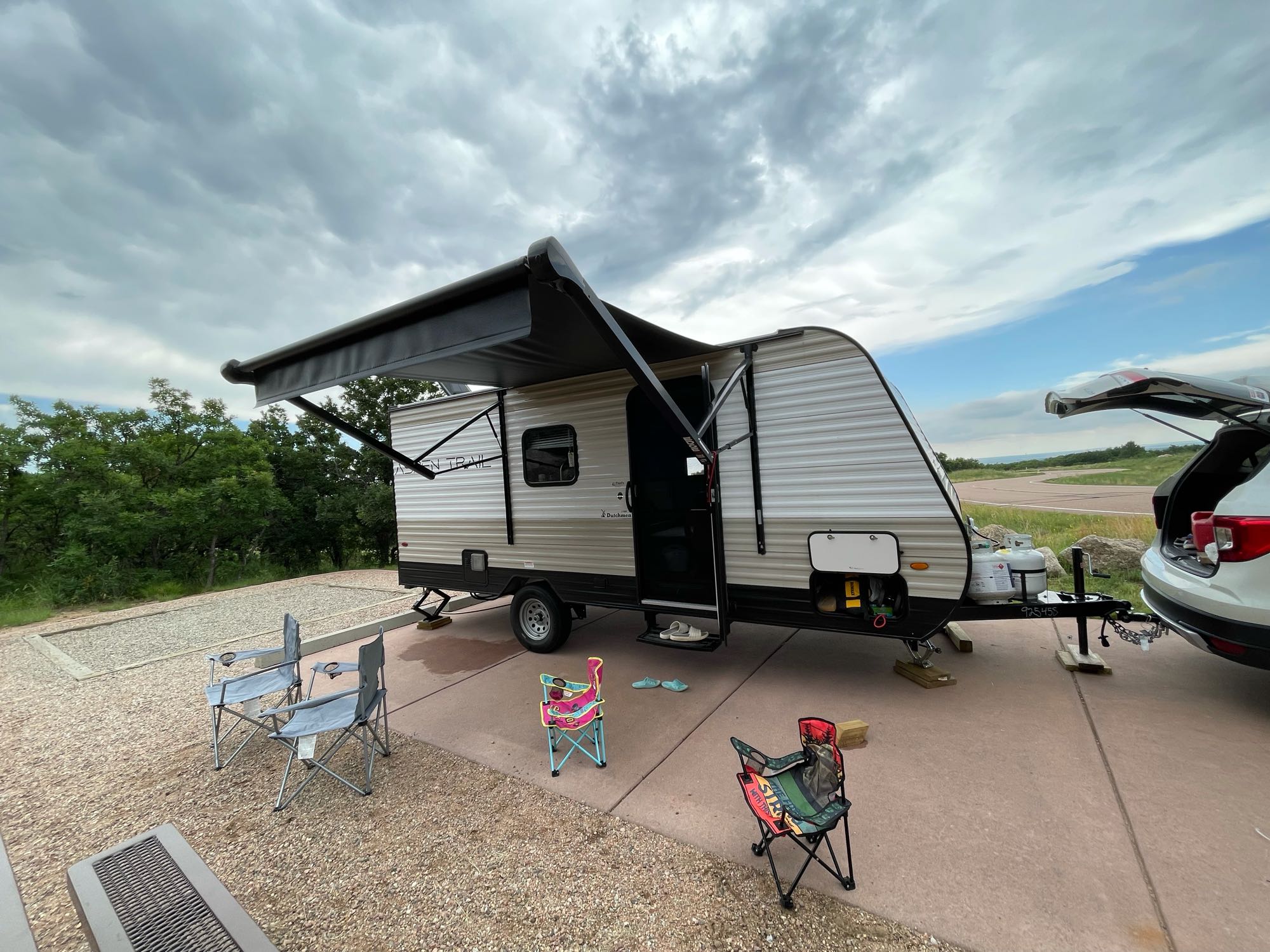 Relaxing RV rental setup at a paved campsite with extended awning, camping chairs, and connected car. Ideal for outdoor adventures.. Dutchmen Aspen Trail 2021