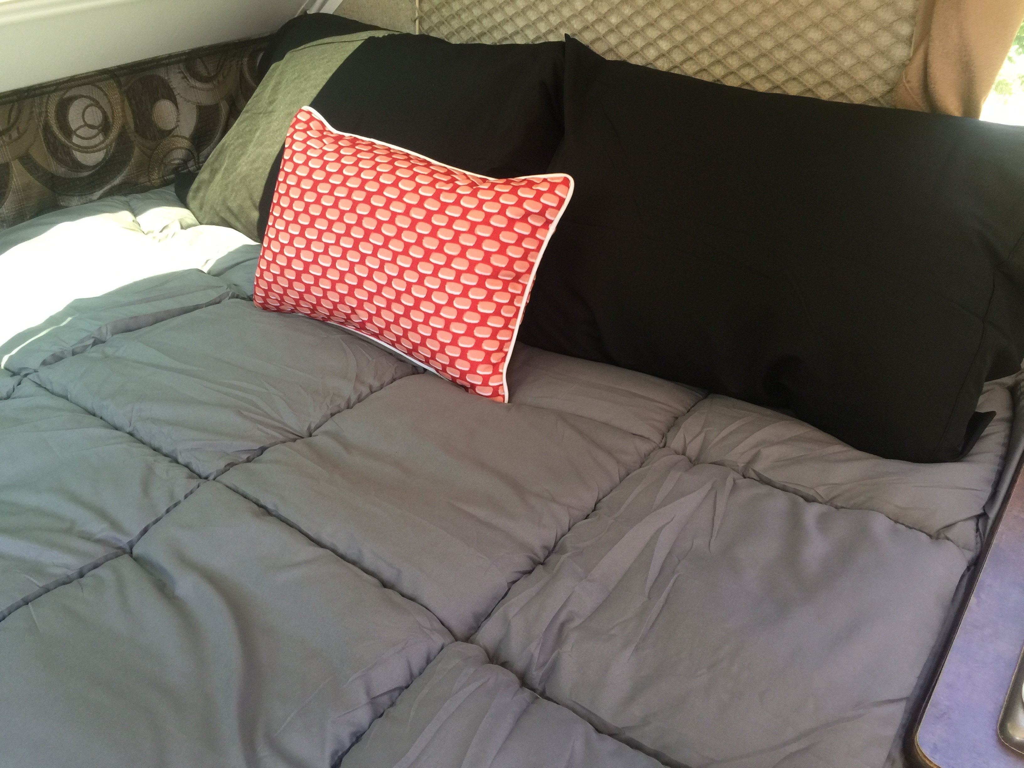 Comfy RV rental bed with gray comforter, dark and red patterned pillows, and patterned interior walls. Bright light.. Forest River Rockwood 2014