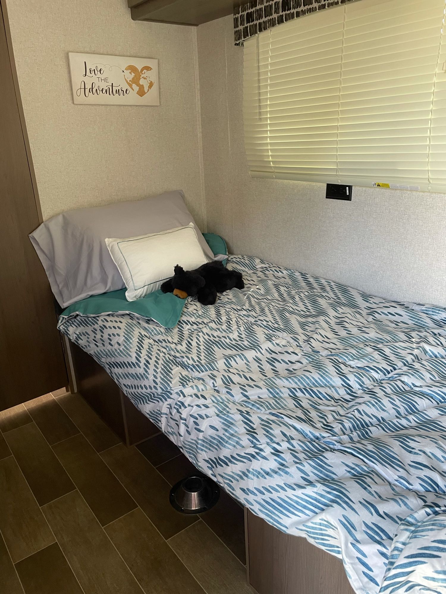 An RV rental's cozy bedroom features a single bed with blue bedding, a "Love the Adventure" sign, and window blinds.. Forest River Independence Trail 2021