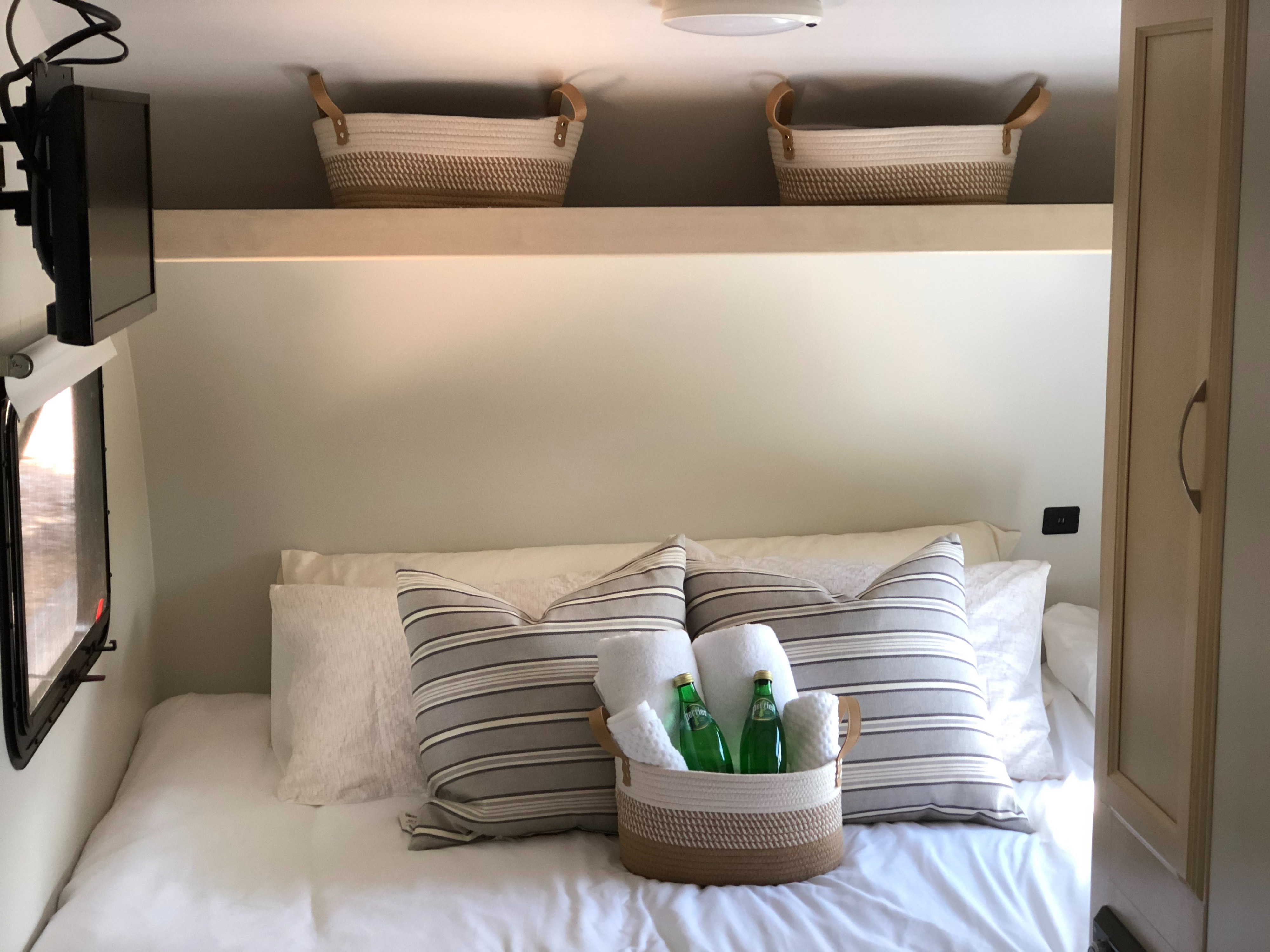 Comfortable RV rental bedroom with white bedding, striped pillows, basket with Perrier/towels, TV, and storage.. Winnebago Minnie Winnie 2018