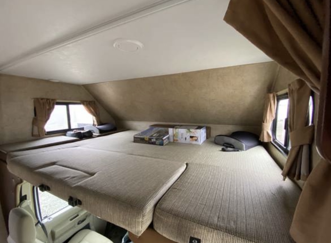 Inside an RV rental: a spacious cab-over bunk bed with a light-colored segmented mattress, windows, and brown curtains.. Forest River Sunseeker 2016