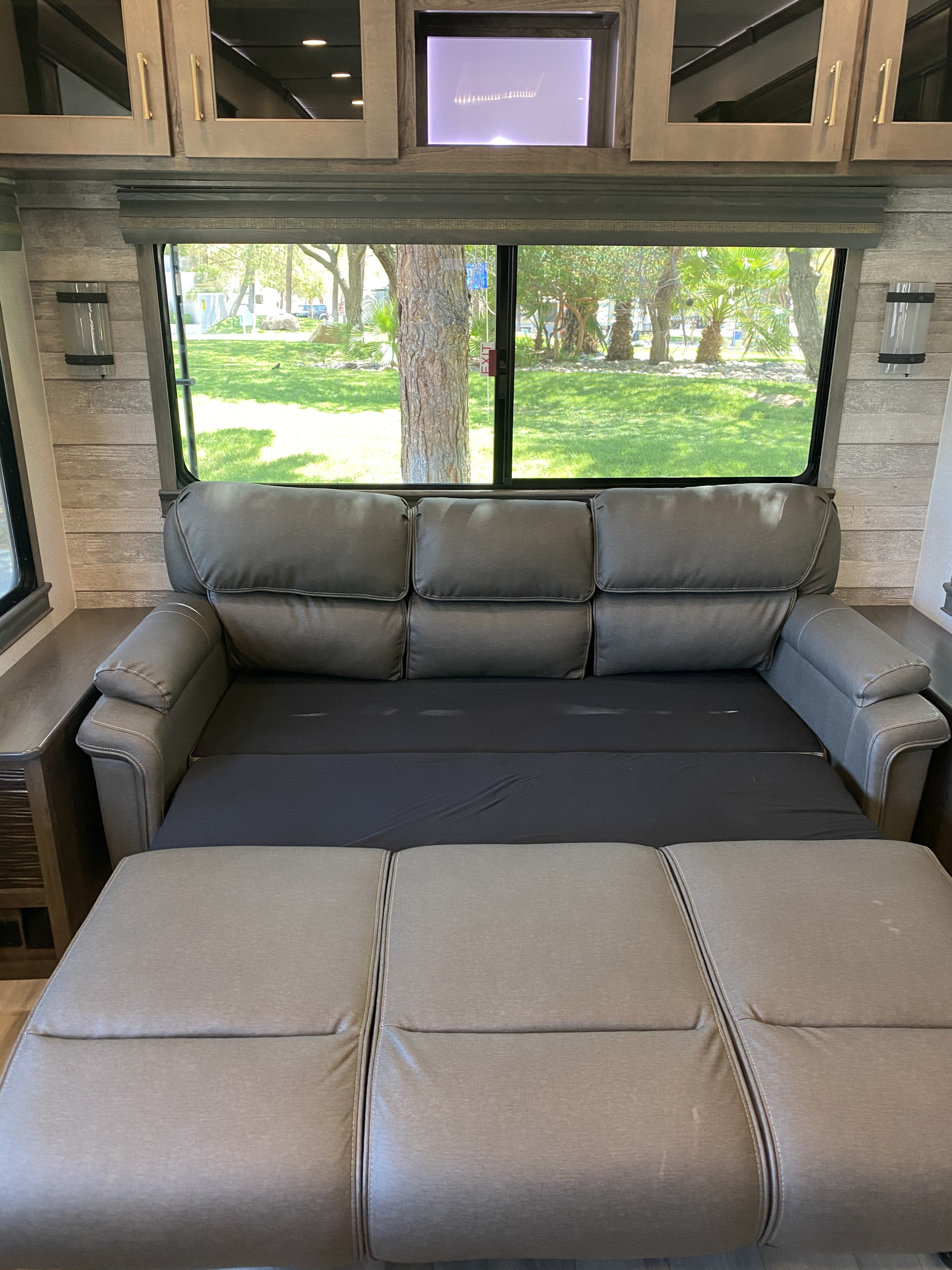 This RV rental's gray sofa bed is extended, showing a comfy sleeping area with a large window view of a green campground.. Alliance RV Paradigm 2021