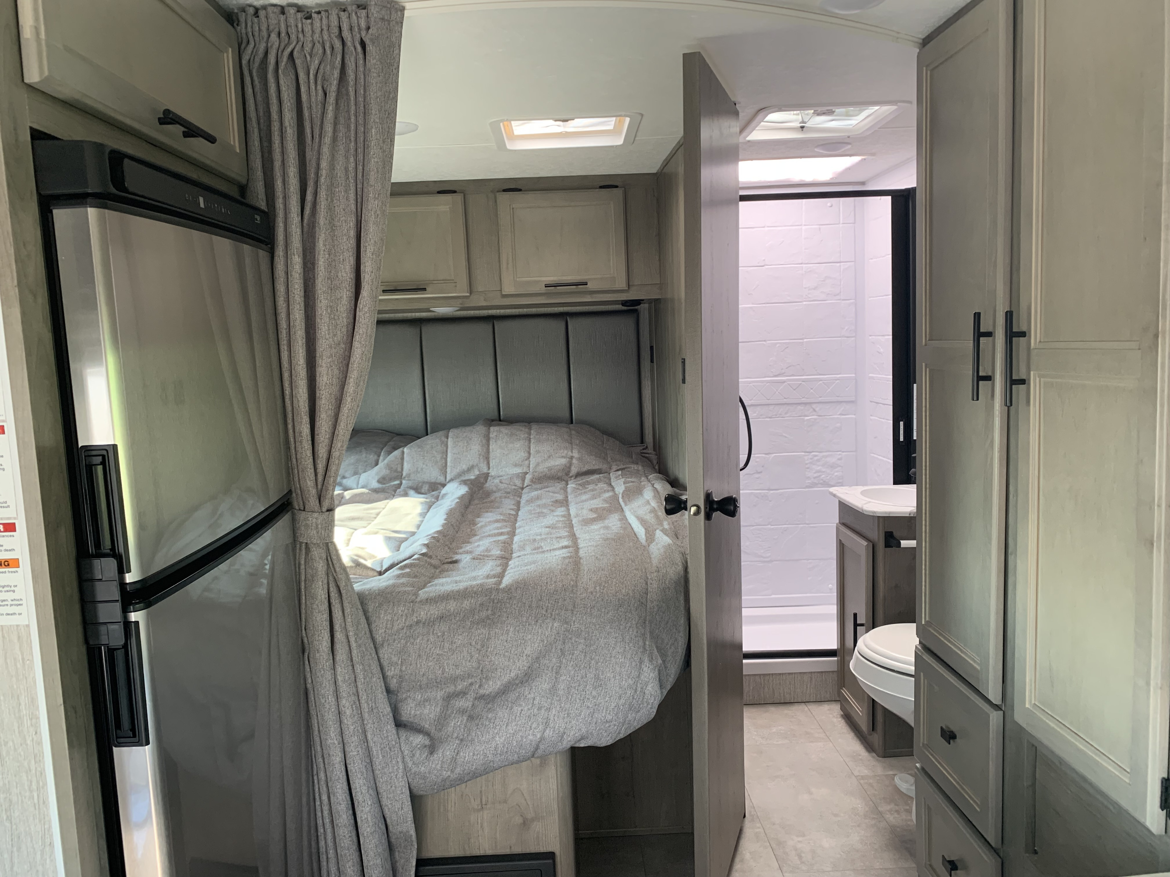 RV rental interior: a comfy bed with overhead cabinets, a refrigerator, and a compact bathroom featuring a shower and toilet.. Other Other 2021
