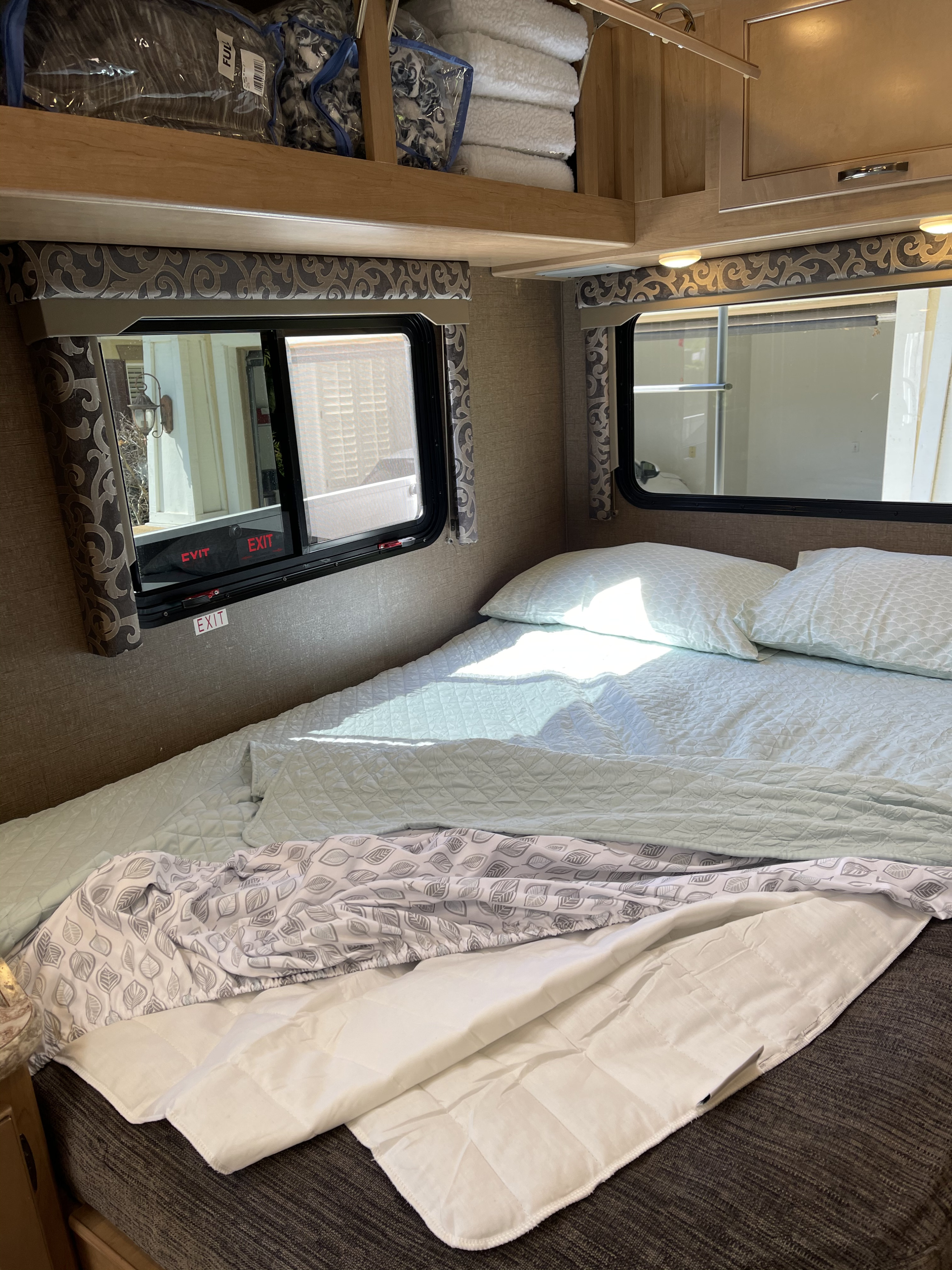 Inside an RV rental: a bright, comfy bedroom with quilted bedding, storage, and an emergency window.. Thor Four Winds 2019