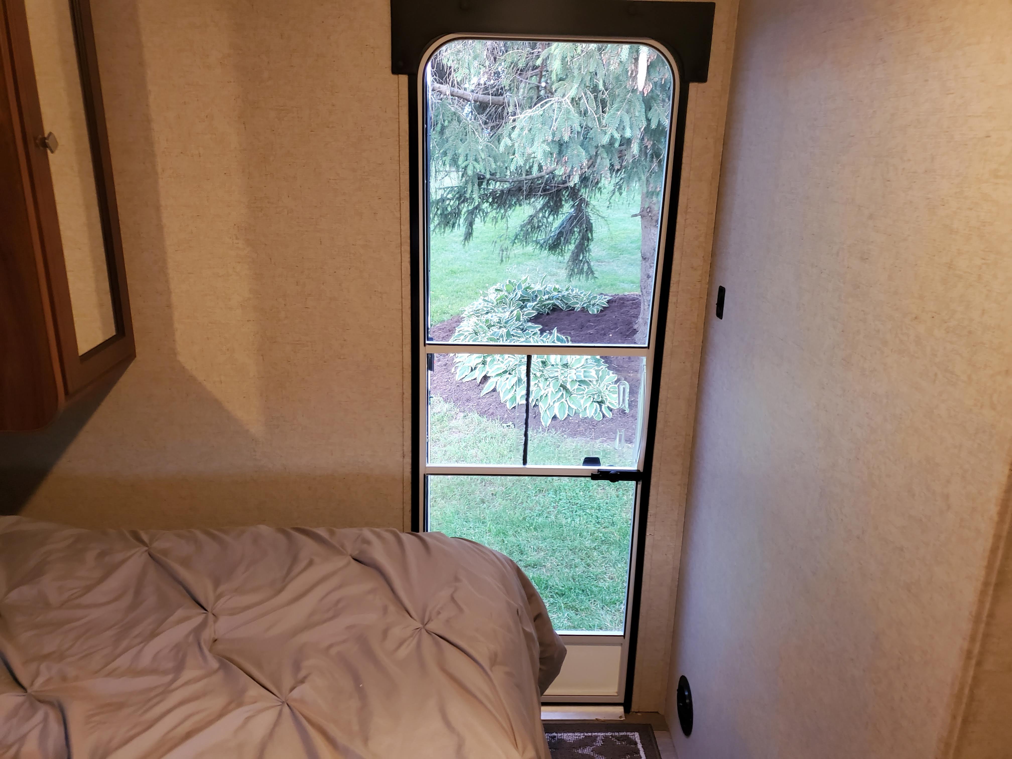 RV rental bedroom: a cozy bed & window offering a tranquil view of green trees & yard.. Heartland Other 2017