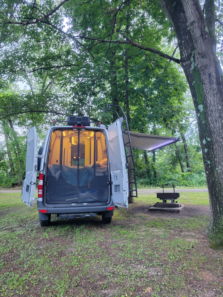 Grey RV rental camper van at a wooded campsite, rear doors open showing the lit interior, with an awning and nearby fire pit.. Winnebago Revel 2019