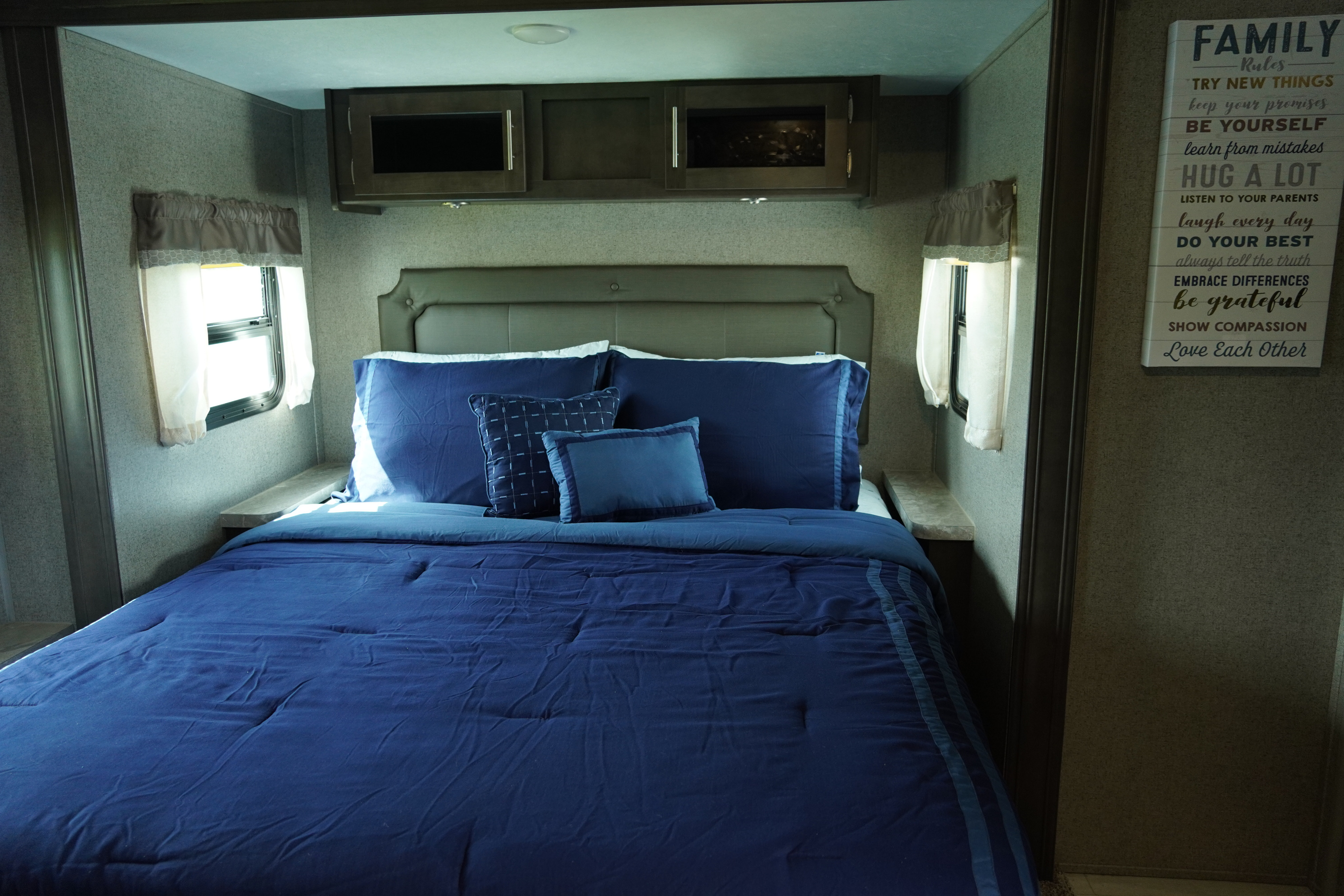 Interior of an RV rental bedroom: cozy blue bed, side windows, overhead cabinets, and a 'Family Rules' wall sign.. Forest River Flagstaff Super Lite 2021