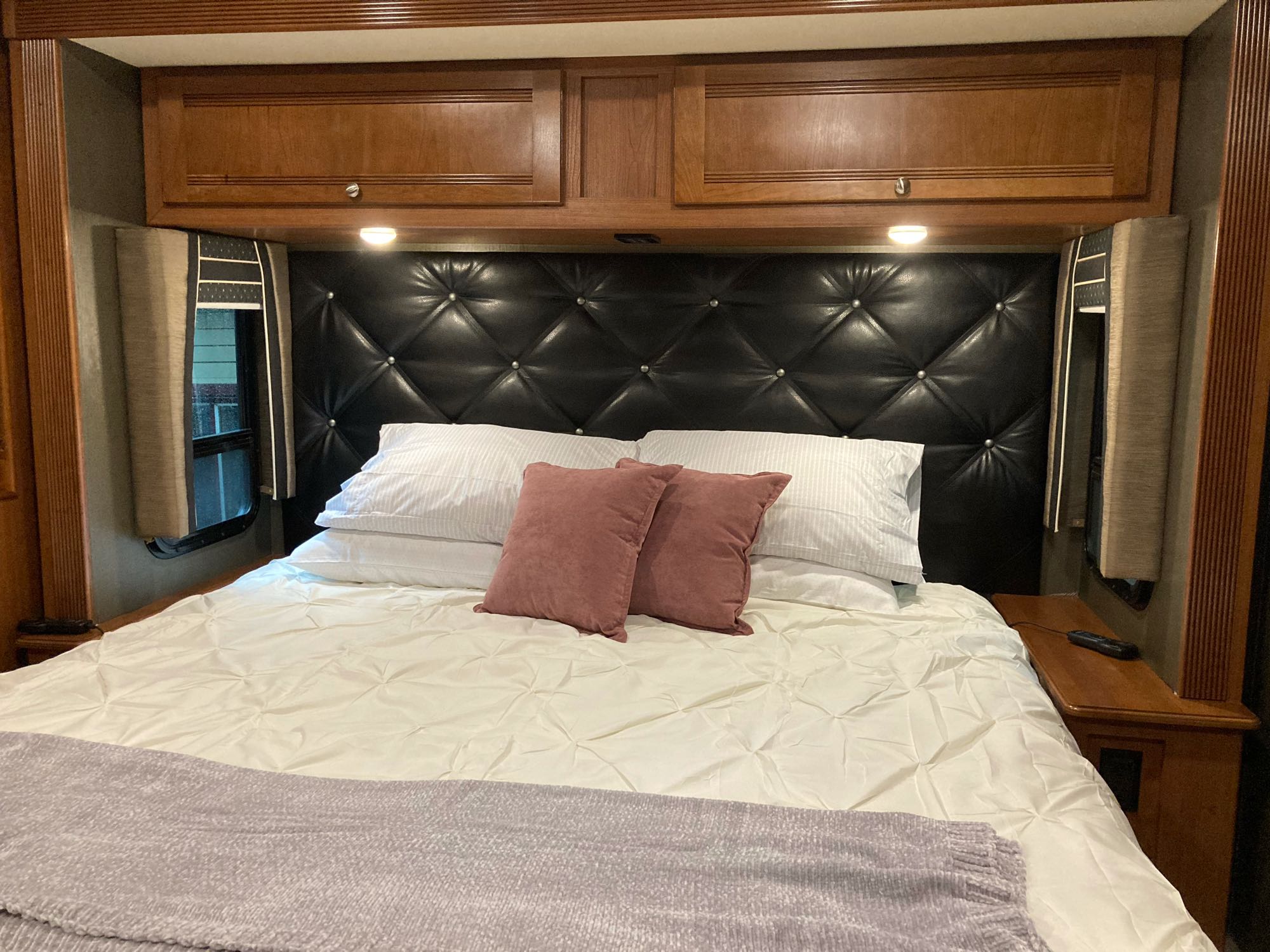 Cozy RV rental bedroom with a black tufted headboard, overhead wooden cabinets, white bedding, and accent pillows.. Winnebago Forza 2018