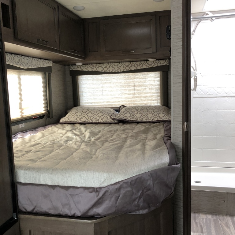 RV rental interior: a bedroom with a bed, overhead cabinets, windows with blinds, and an adjacent shower.. Thor Motor Coach Freedom Elite 2021