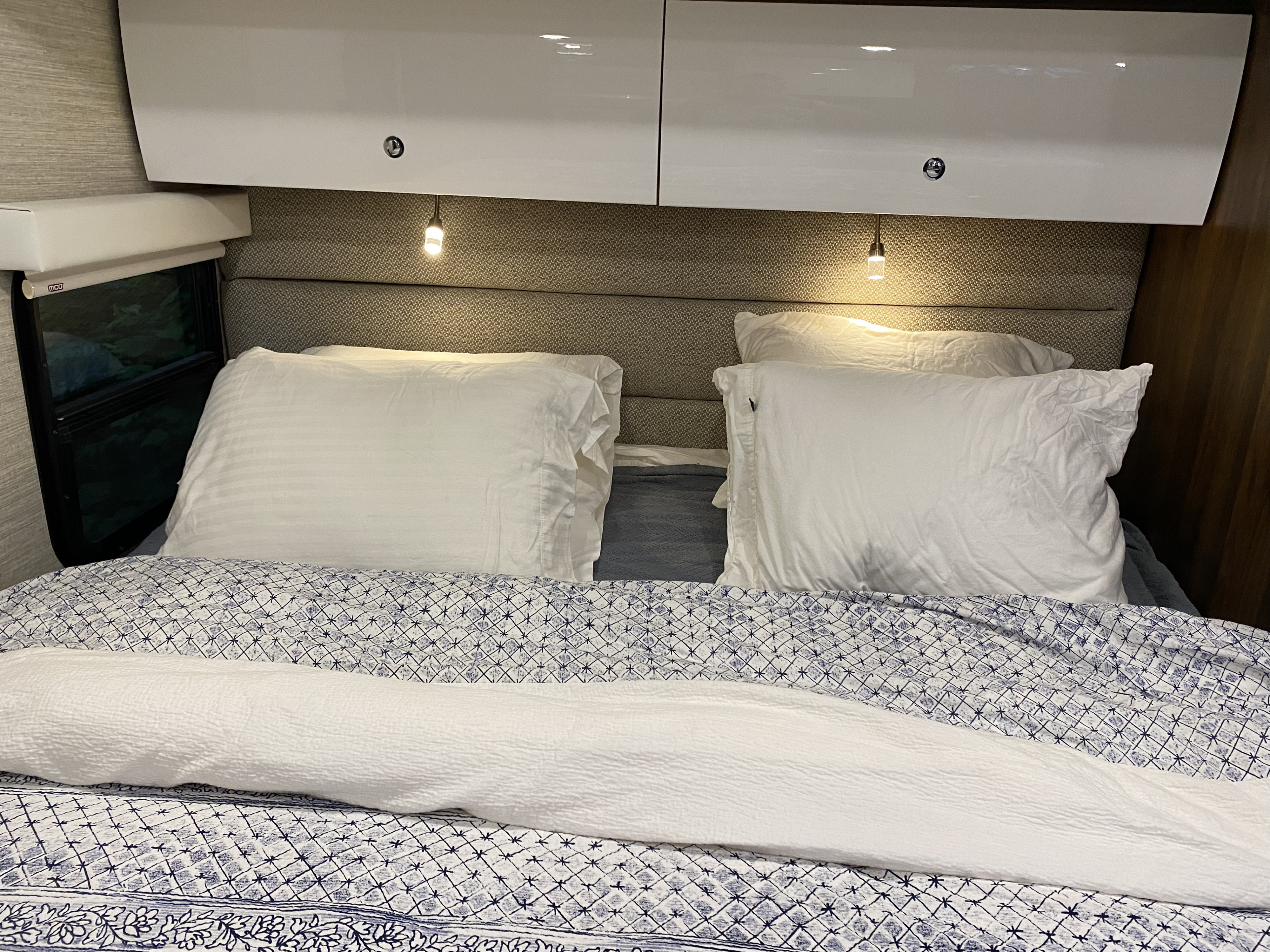 Cozy RV rental bedroom featuring a bed with white pillows, blue patterned bedding, overhead cabinets, and reading lights.. Winnebago View 2021