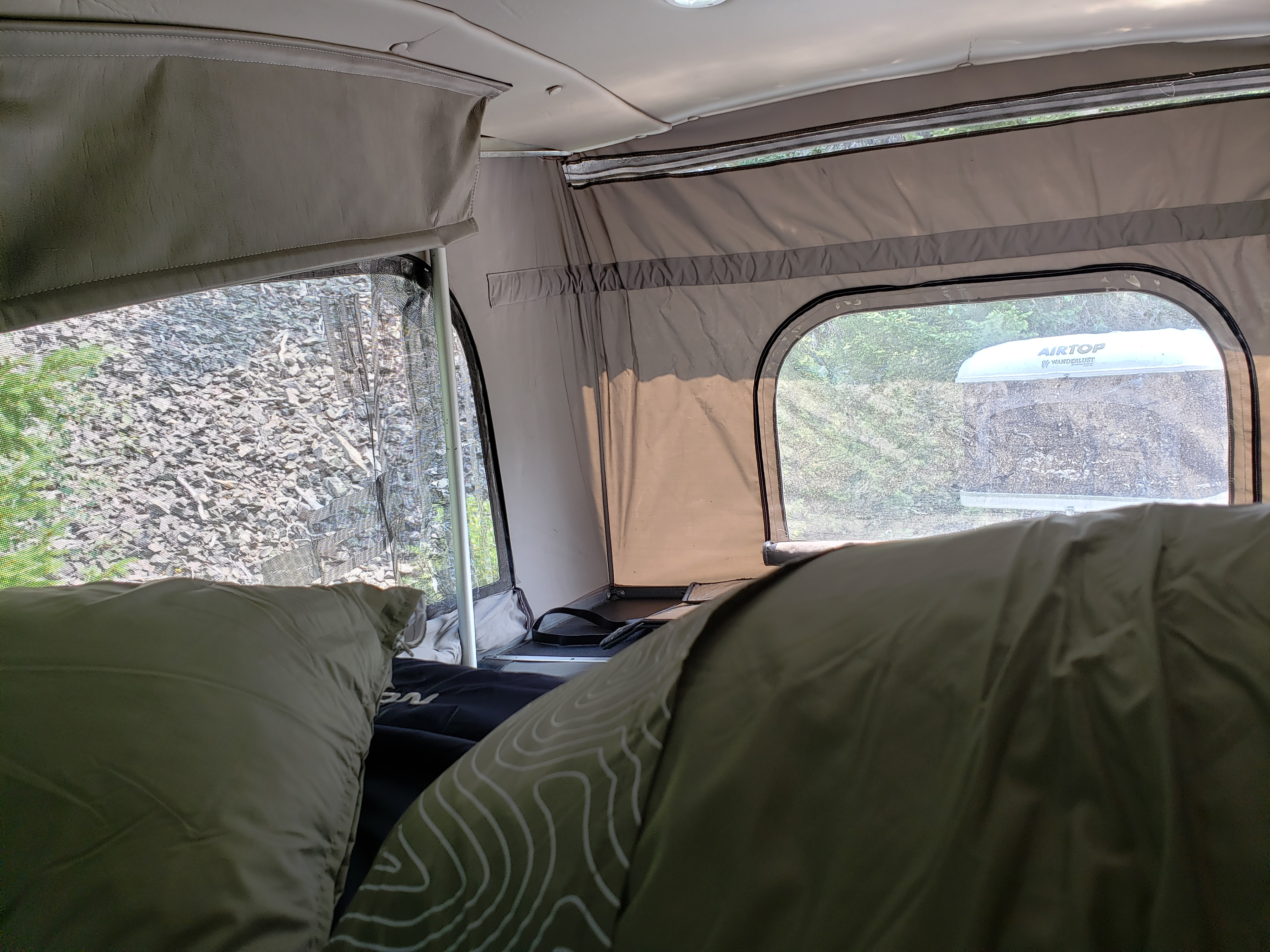 Inside an RV rental, cozy bedding offers views through windows of a gravel area and an 'AIRTOP' camper top outside.. Roadtrek N6-Active 2012