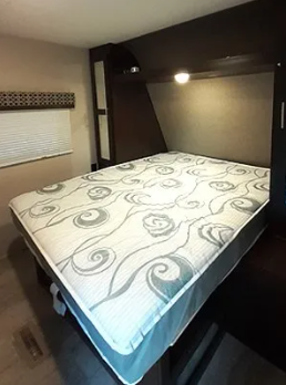 Inside an RV rental, a comfy queen bed with a patterned mattress cover, overhead storage, and reading lights.. KZSP Sportsman 260 2019