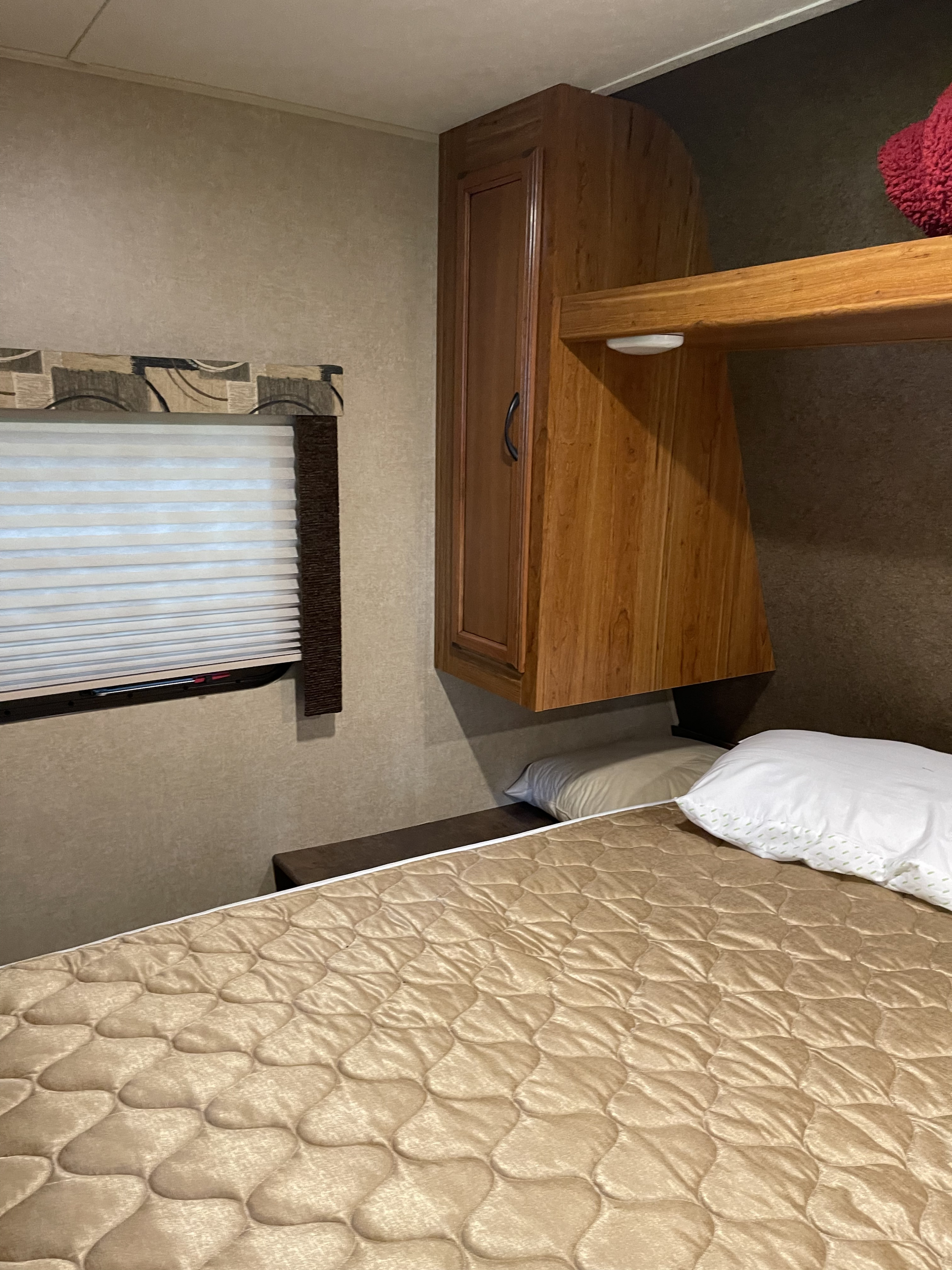 Compact RV rental bedroom featuring a quilted bed, pillows, wooden overhead cabinets, and a window with pleated shades.. Starcraft Launch 2016