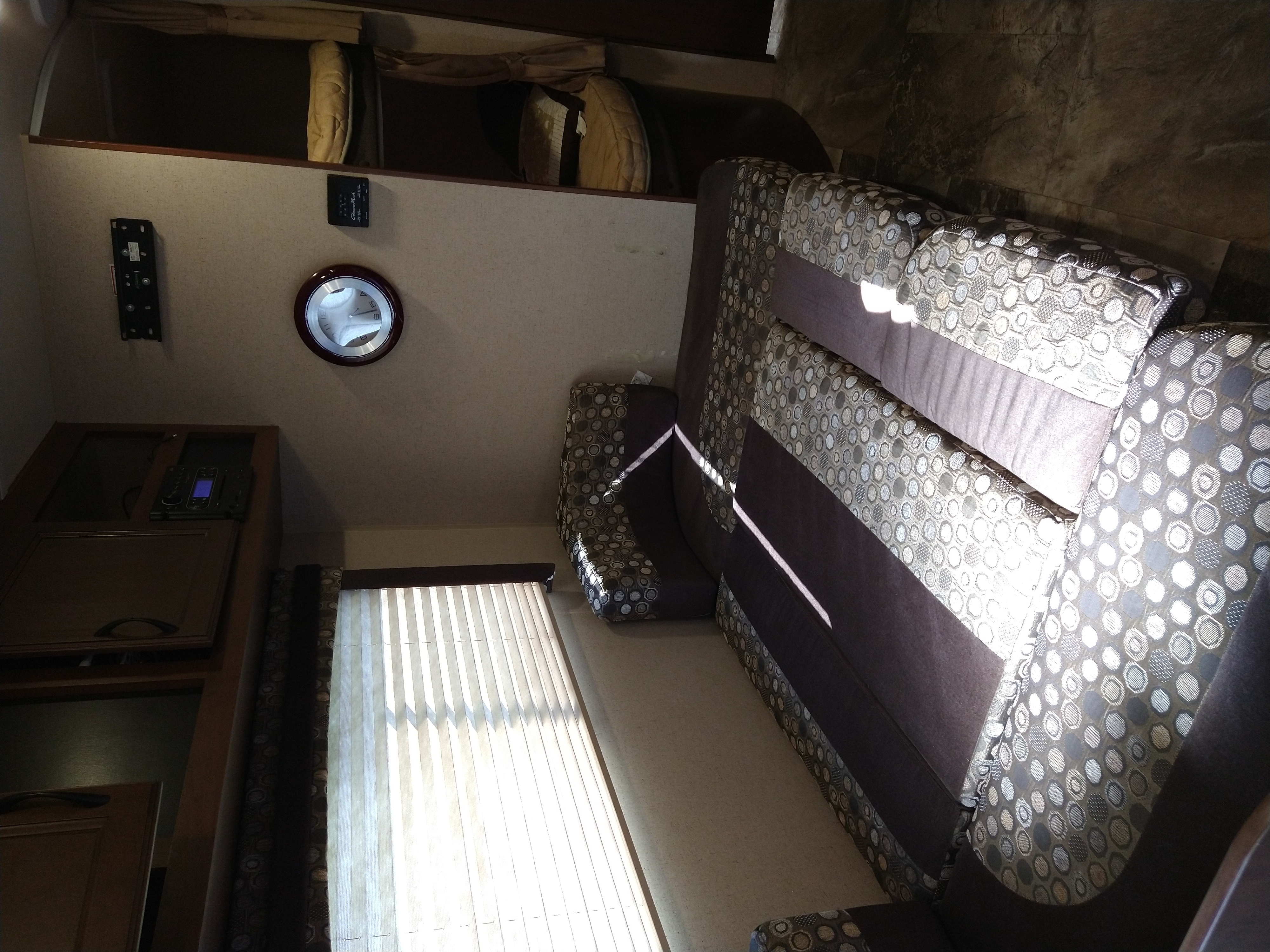 Interior of an RV rental showing a patterned sofa, sunlit window with blinds, an entertainment console, and overhead bunk.. Jayco Jay Flight 2015