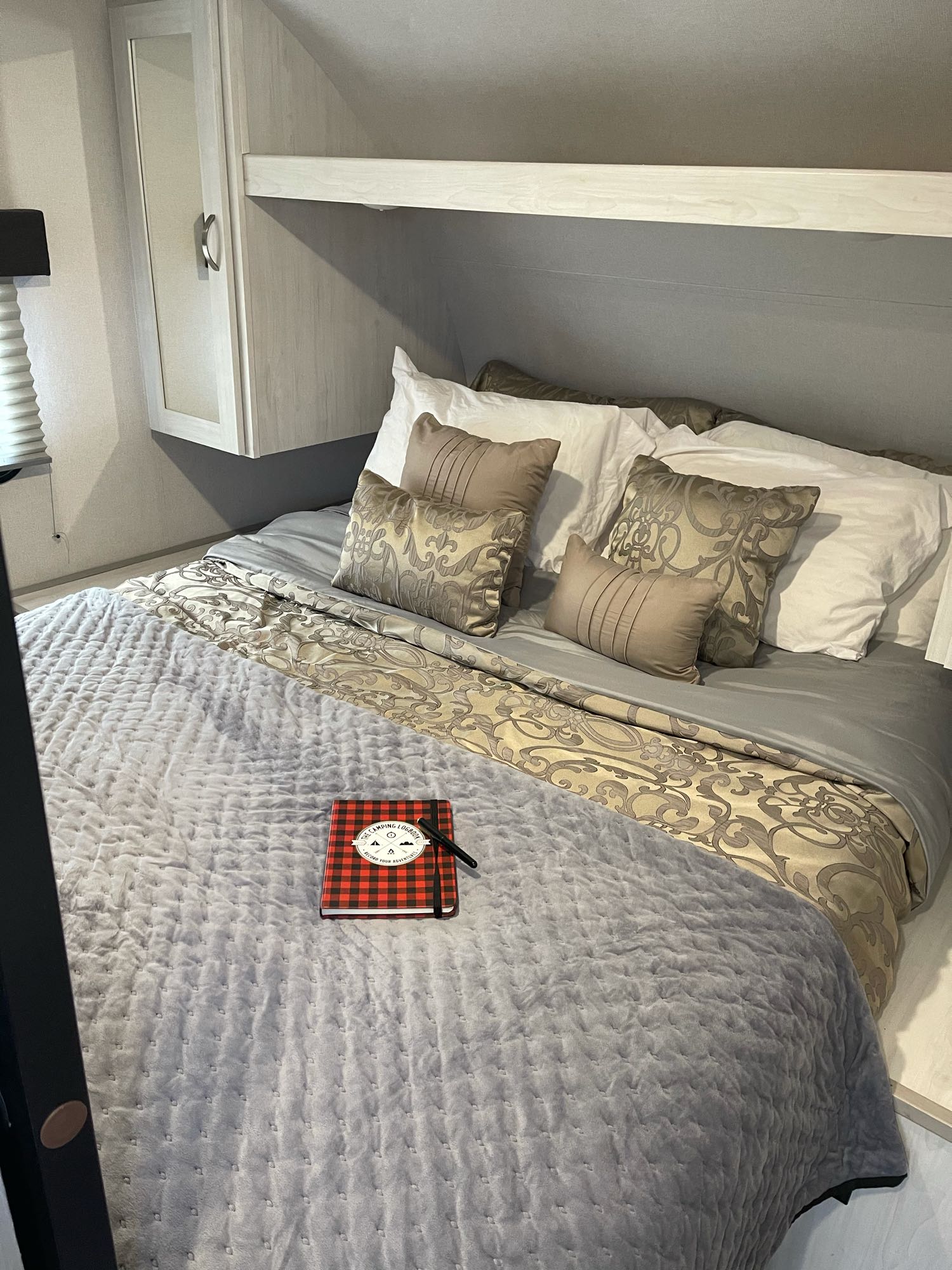 Comfortable RV rental bed with soft gray quilt, decorative pillows, and a camping logbook on top. Built-in storage above.. Prime Time Tracer 2021