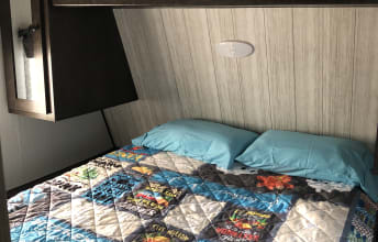This RV rental bedroom features a comfy bed with a colorful quilt and blue pillows, perfect for a relaxing travel trailer stay.. Heartland Pioneer 2020