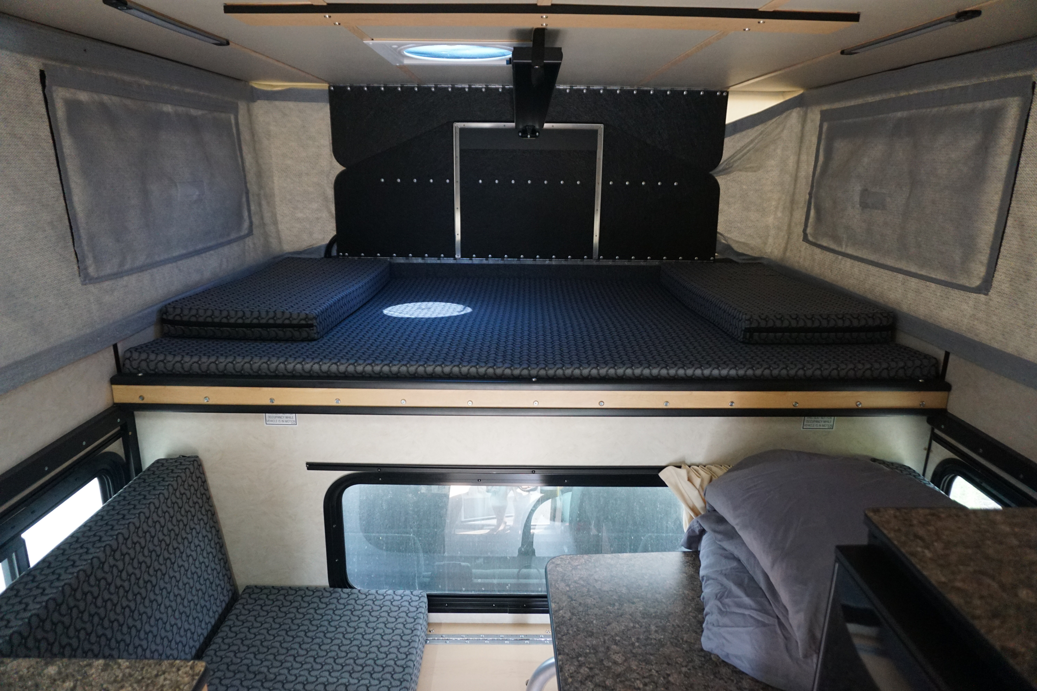 RV rental interior: A bed/lounge area with patterned gray cushions and a dining table on the right.. Chevrolet Silverado 2020