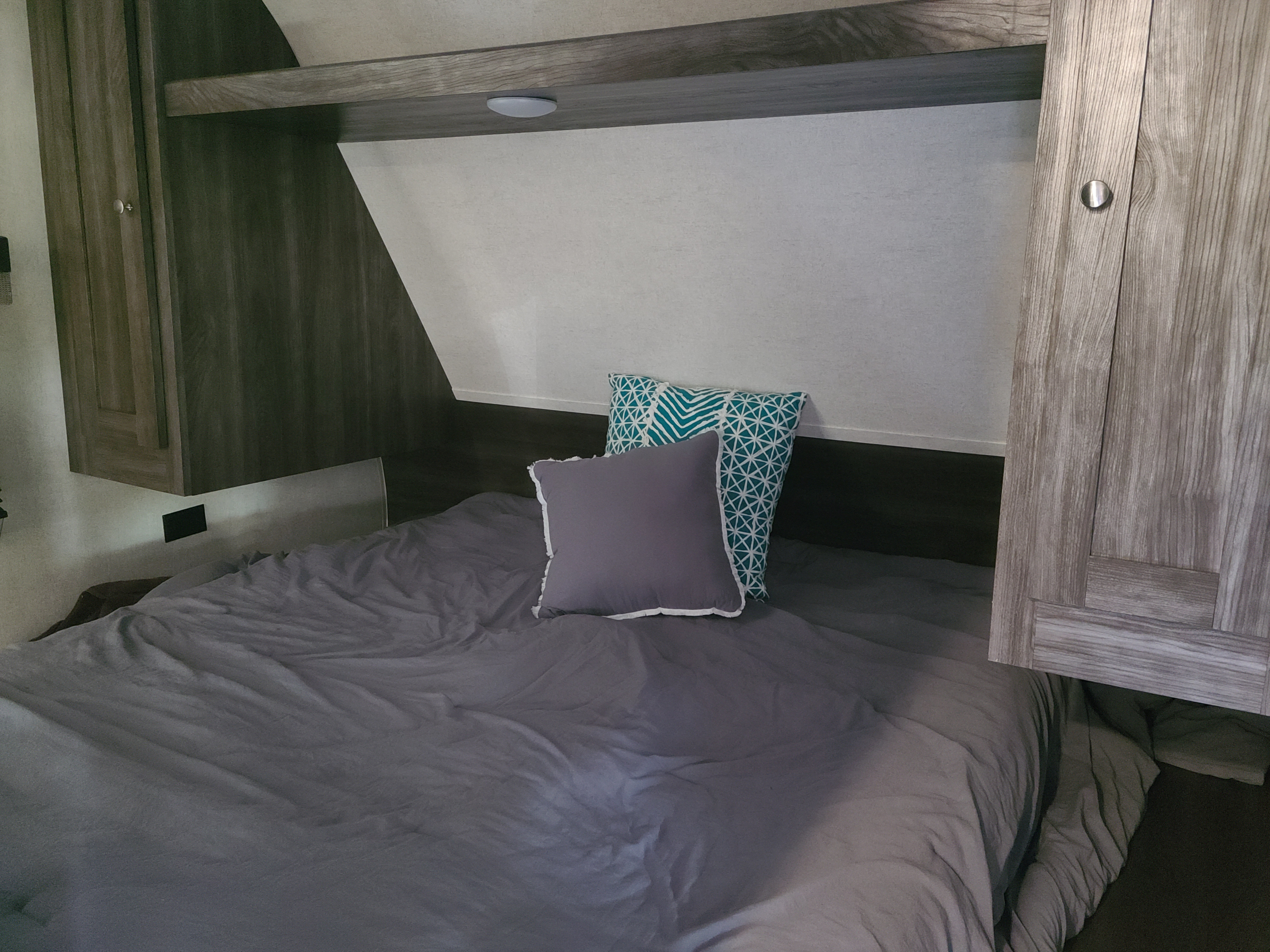 An RV rental's inviting bedroom area featuring a gray bed, patterned pillows, overhead shelf with light, and wooden cabinets.. Open Range Open Range 2019