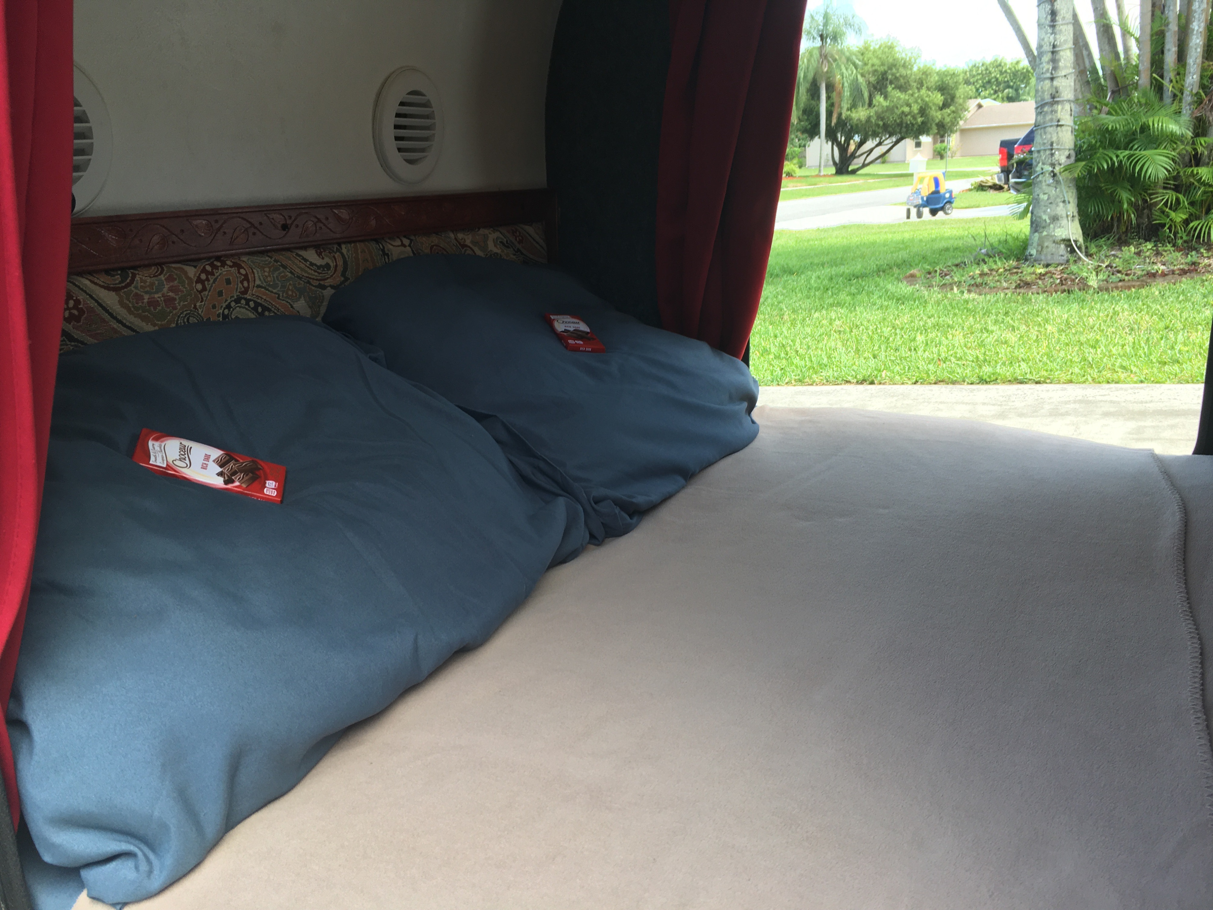 Inside an RV rental: a cozy bed with blue pillows and chocolate bars, red curtains opening to a grassy street with a toy car.. Little Guy Teardrop Trailer 2007