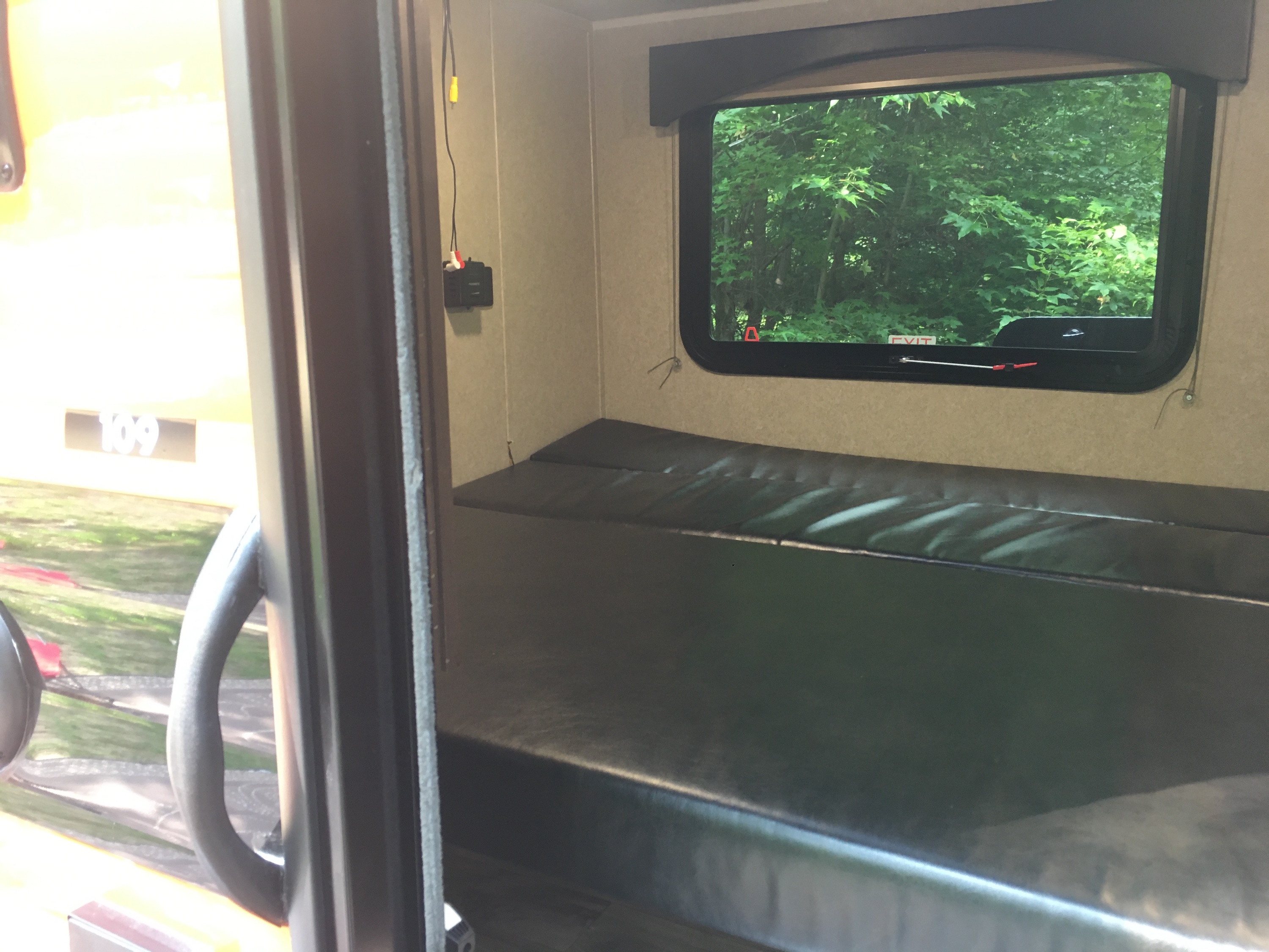 Inside an RV rental, a dark bunk bed faces a window with lush green trees. An exit sign and wall wiring are visible.. Sunset Park Sunray 109 2021