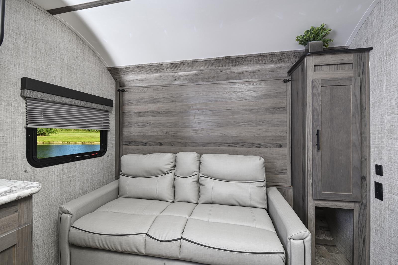 Inside an RV rental: a modern, light-grey sofa, window with a scenic lake view, and chic wooden wall paneling. Cozy living space.. Gulf Stream Ameri-Lite 2021