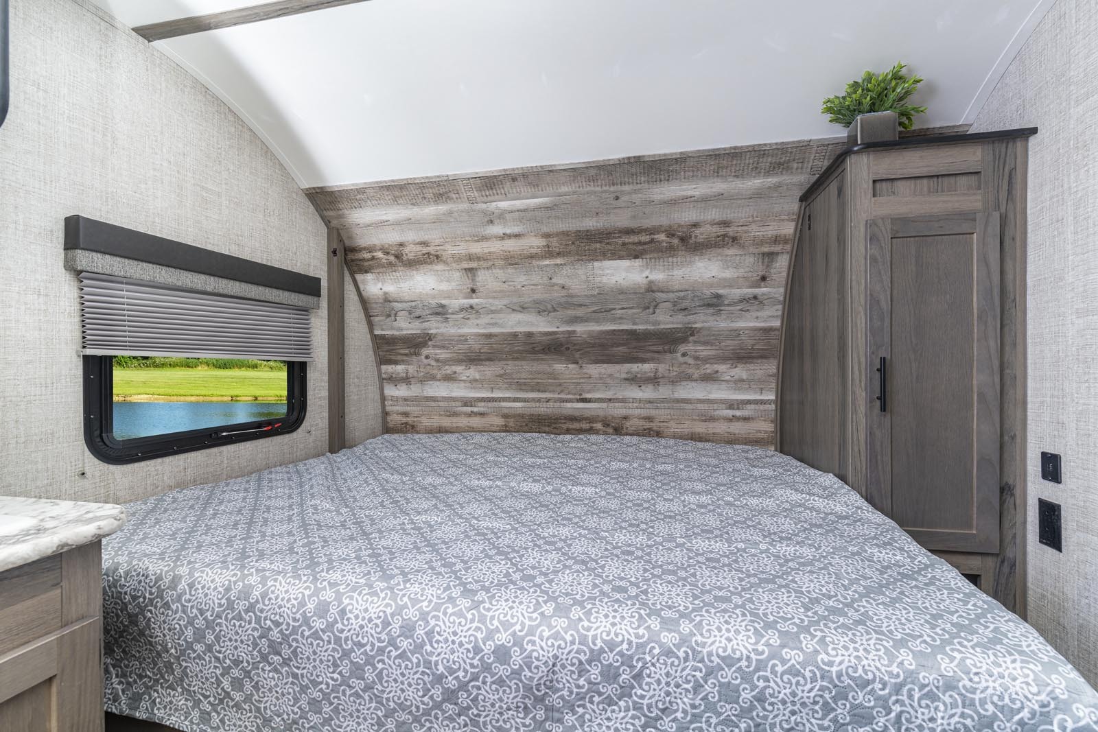 Cozy RV rental bedroom with a patterned bed, rustic wood headboard, and a window showcasing a serene lake view. Perfect for trips.. Gulf Stream Ameri-Lite 2021
