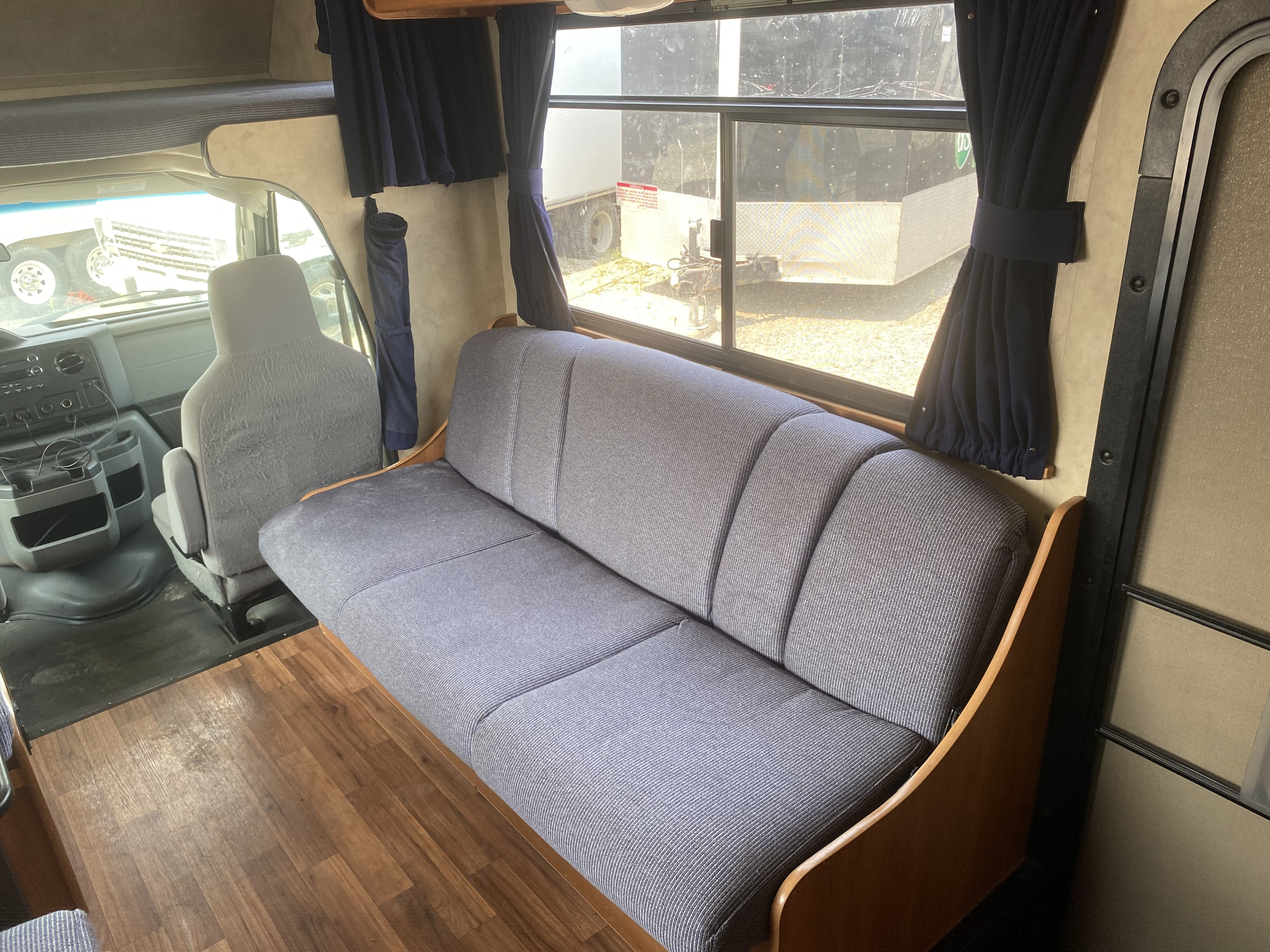 RV rental interior featuring a cozy gray sofa, large window with blue curtains, and a view of the driver's area with wood-look flooring.. Thor Motor Coach Four Winds Majestic 2012