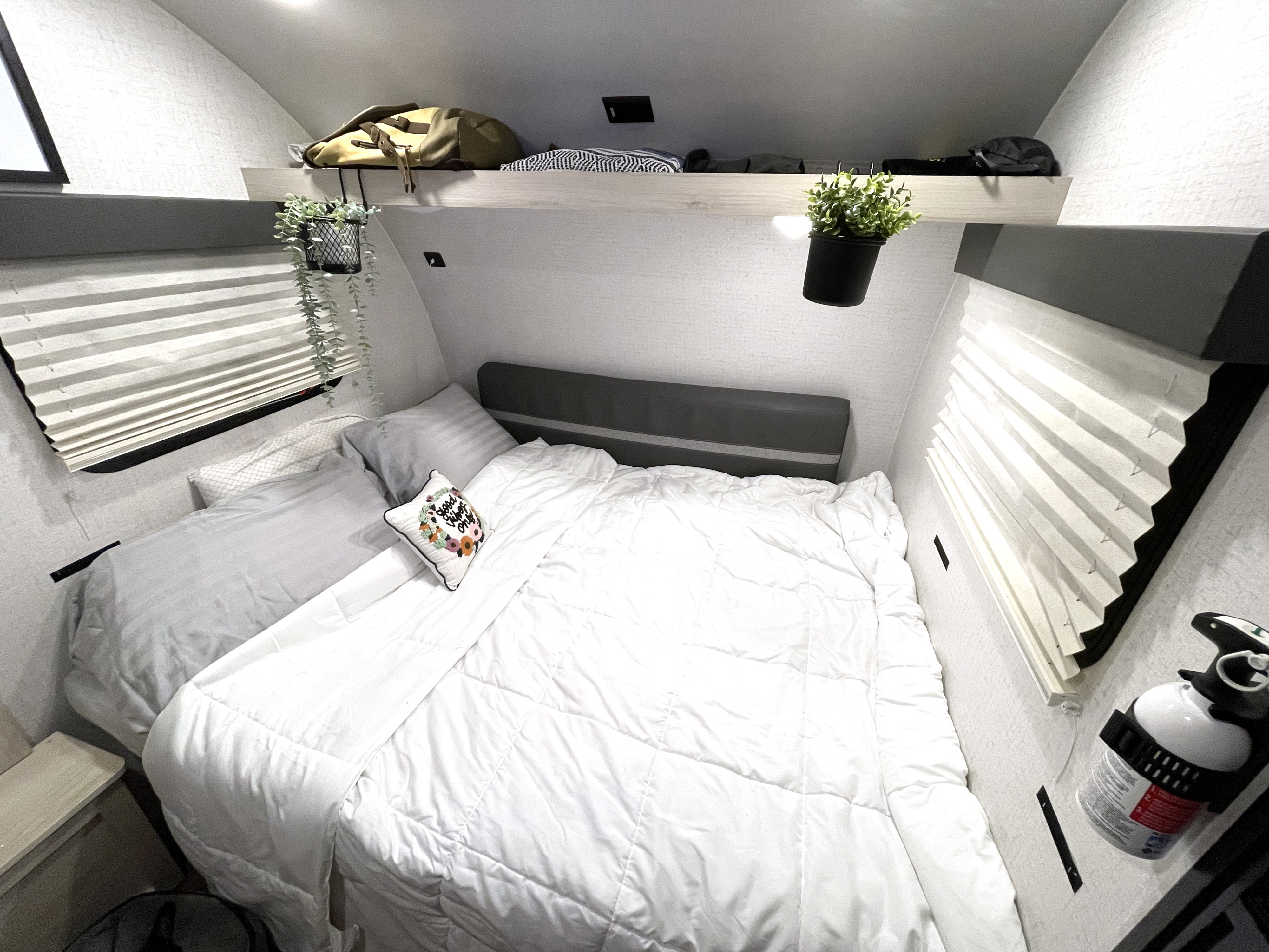 Relaxing RV rental bedroom with a plush white comforter, overhead shelving, pleated window blinds, and charming decor.. Winnebago Hike 2021