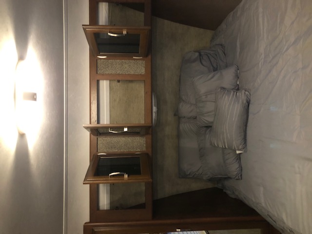 RV rental bedroom with gray bedding, a modern wall light, and wood overhead storage cabinets.. Keystone Passport 2015