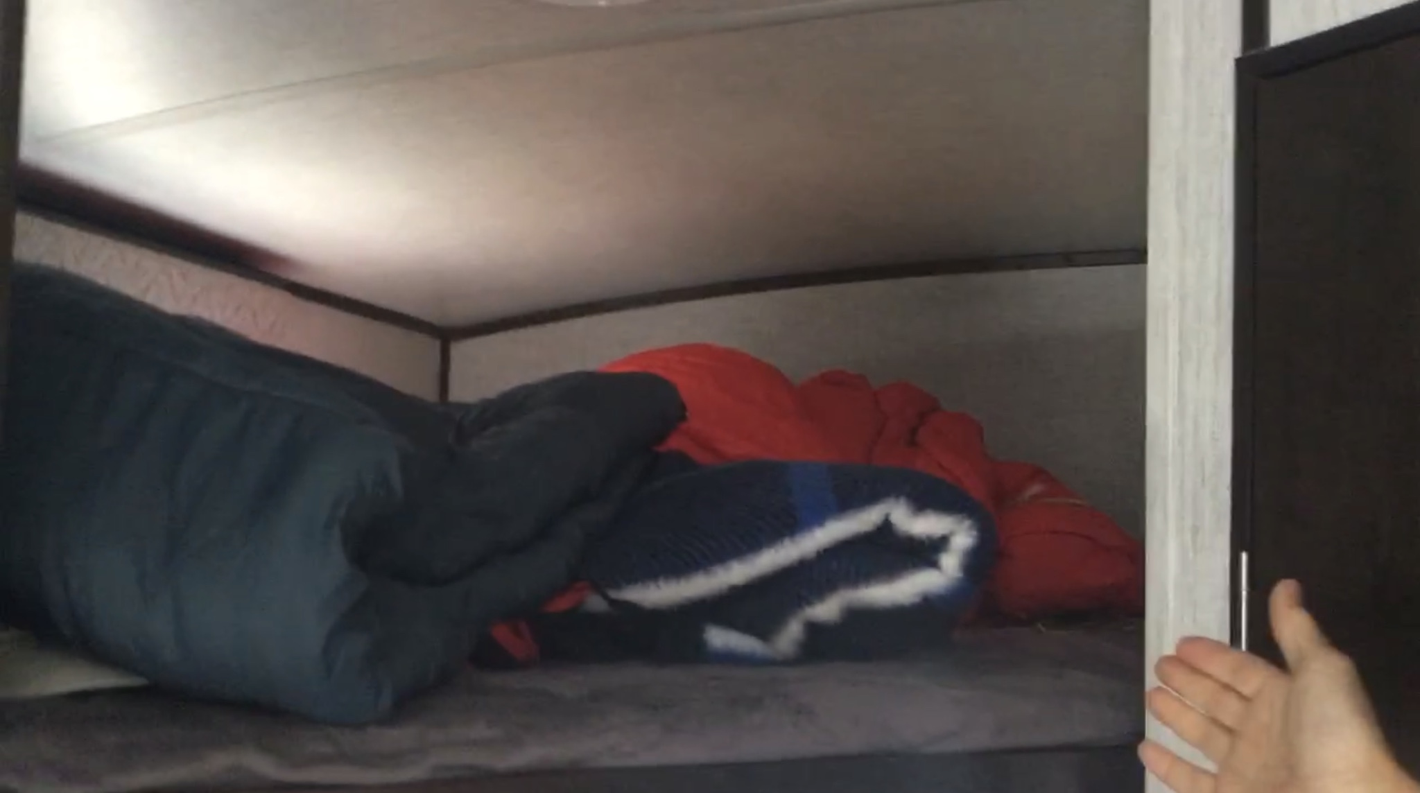An RV rental bunk bed is seen from inside, filled with a blue sleeping bag, orange and striped blankets, and grey bedding.. Heartland RVs Sundance Ultra Lite 2020