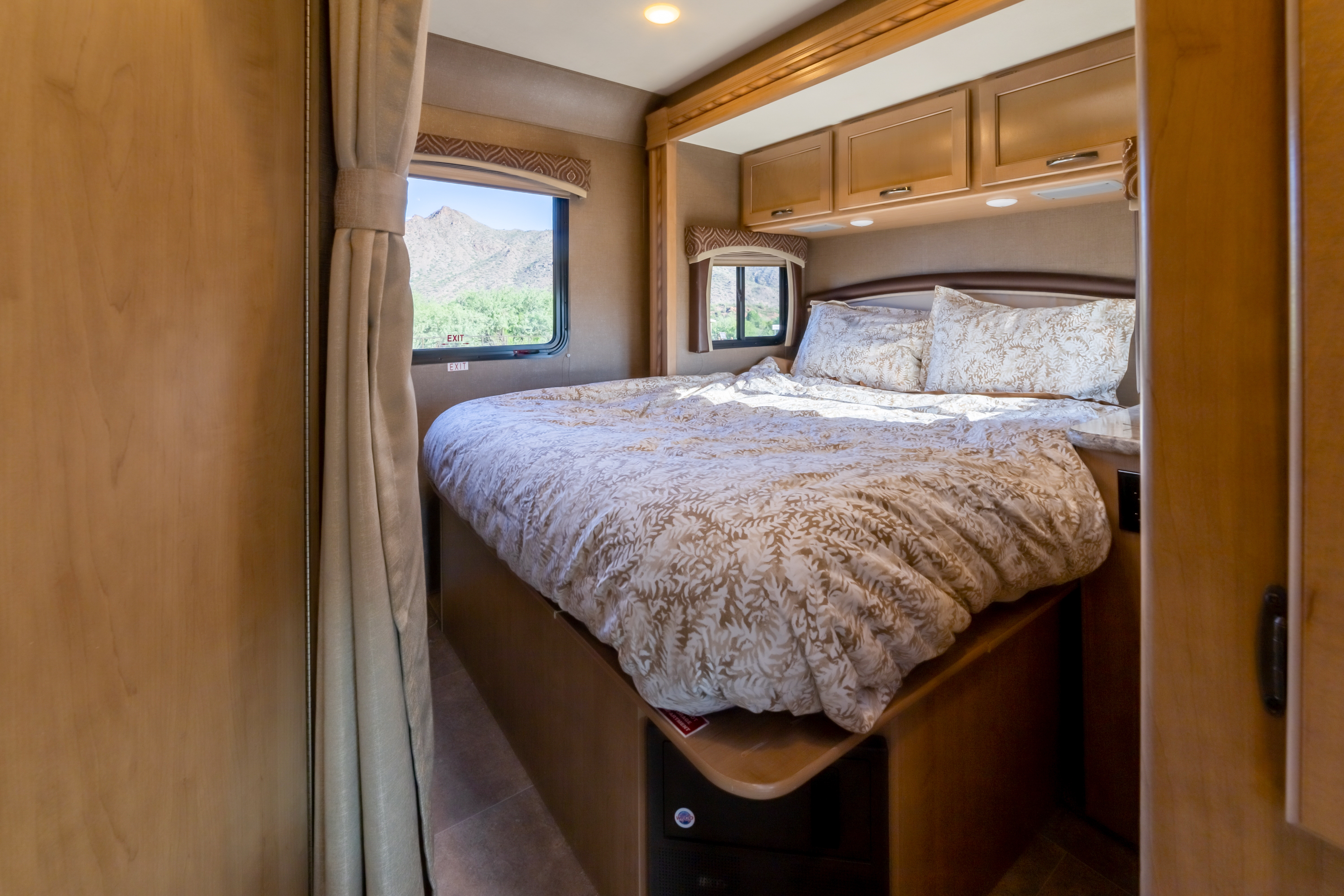 Inside an RV rental, a cozy bedroom features a patterned bed, wood cabinetry, and a scenic mountain view from the window.. Thor Motor Coach Chateau 2018