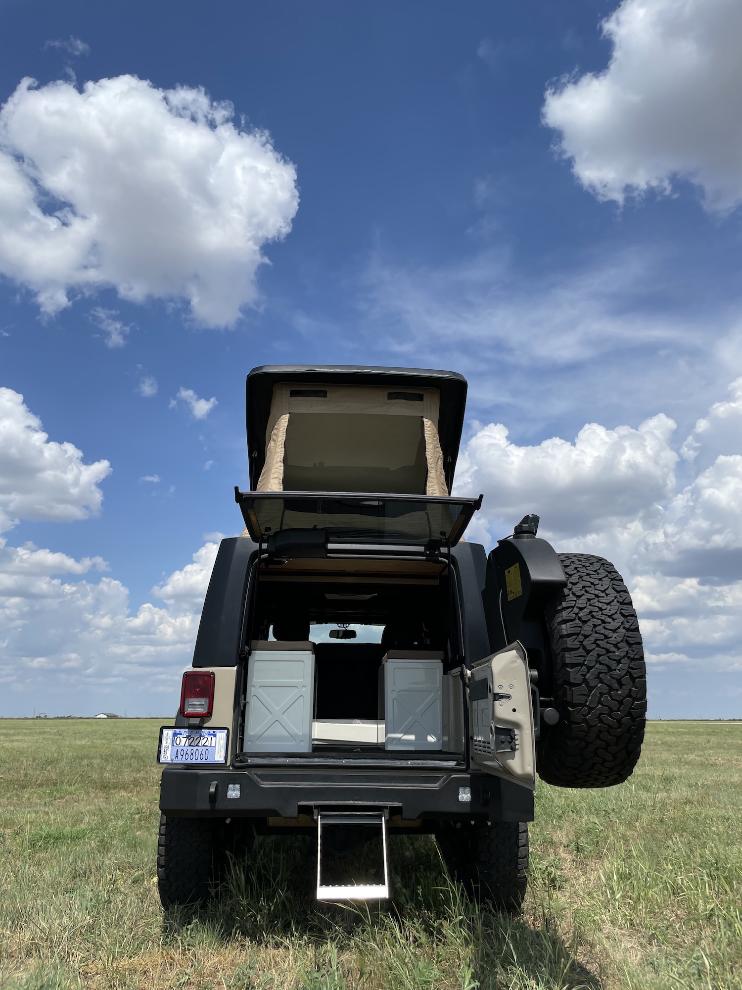 Adventure-ready RV rental: A tan Jeep with a raised pop-top camper and open tailgate revealing gear, parked in a sunny, grassy field.. Jeep Rubicon Pop-top 2016