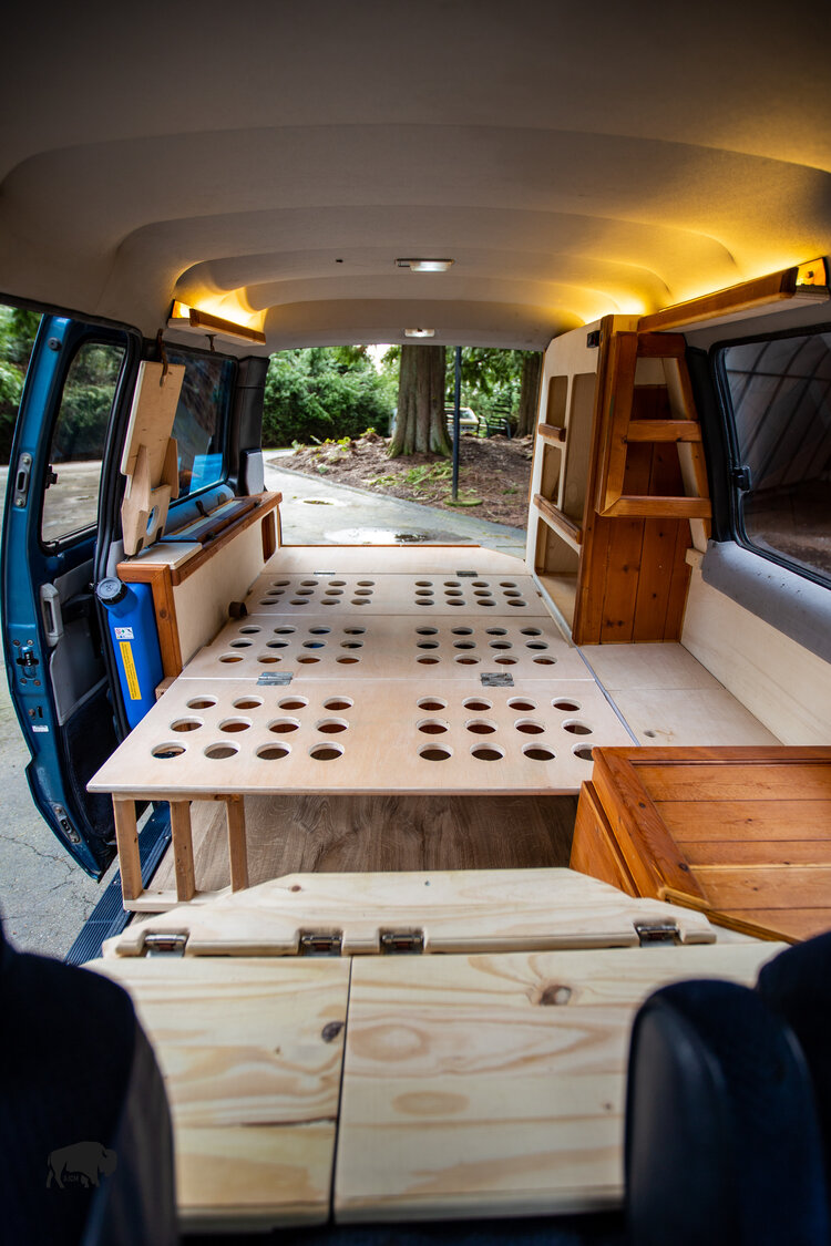 This RV rental's versatile interior: a custom van with wooden convertible bed, built-in storage, and warm lighting.. Toyota Hiace 1985