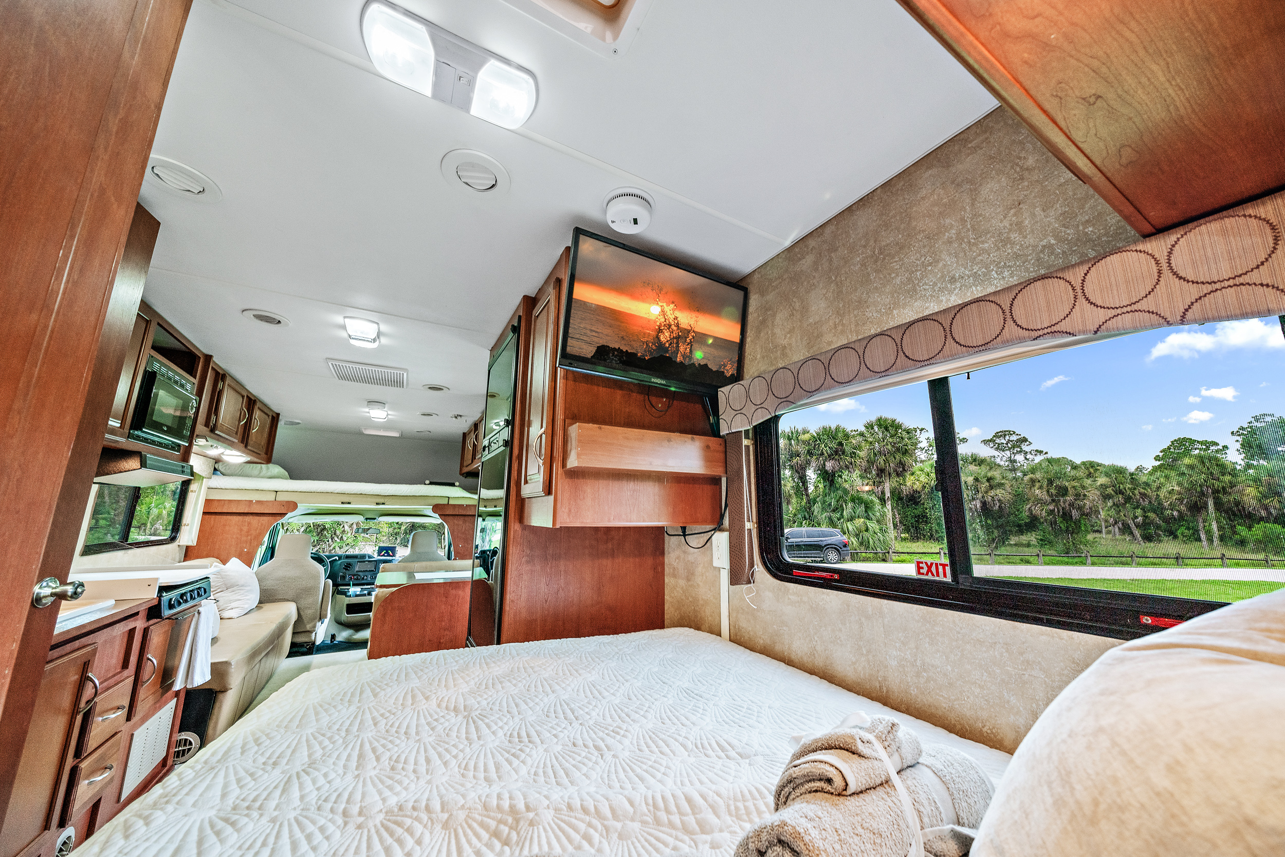 Spacious rv rental interior featuring a cozy bed, TV, kitchen, and a window view of palm trees outside.. Winnebago Spirit 2015
