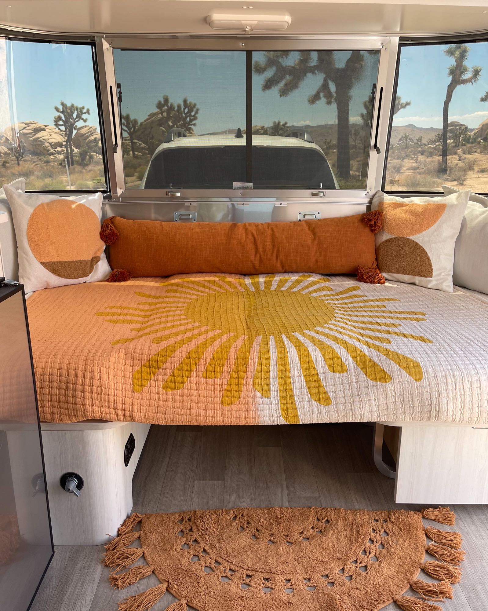 Cozy RV rental interior with a sun-patterned bed and pillows, overlooking a desert landscape with Joshua trees.. Airstream Bambi 2021