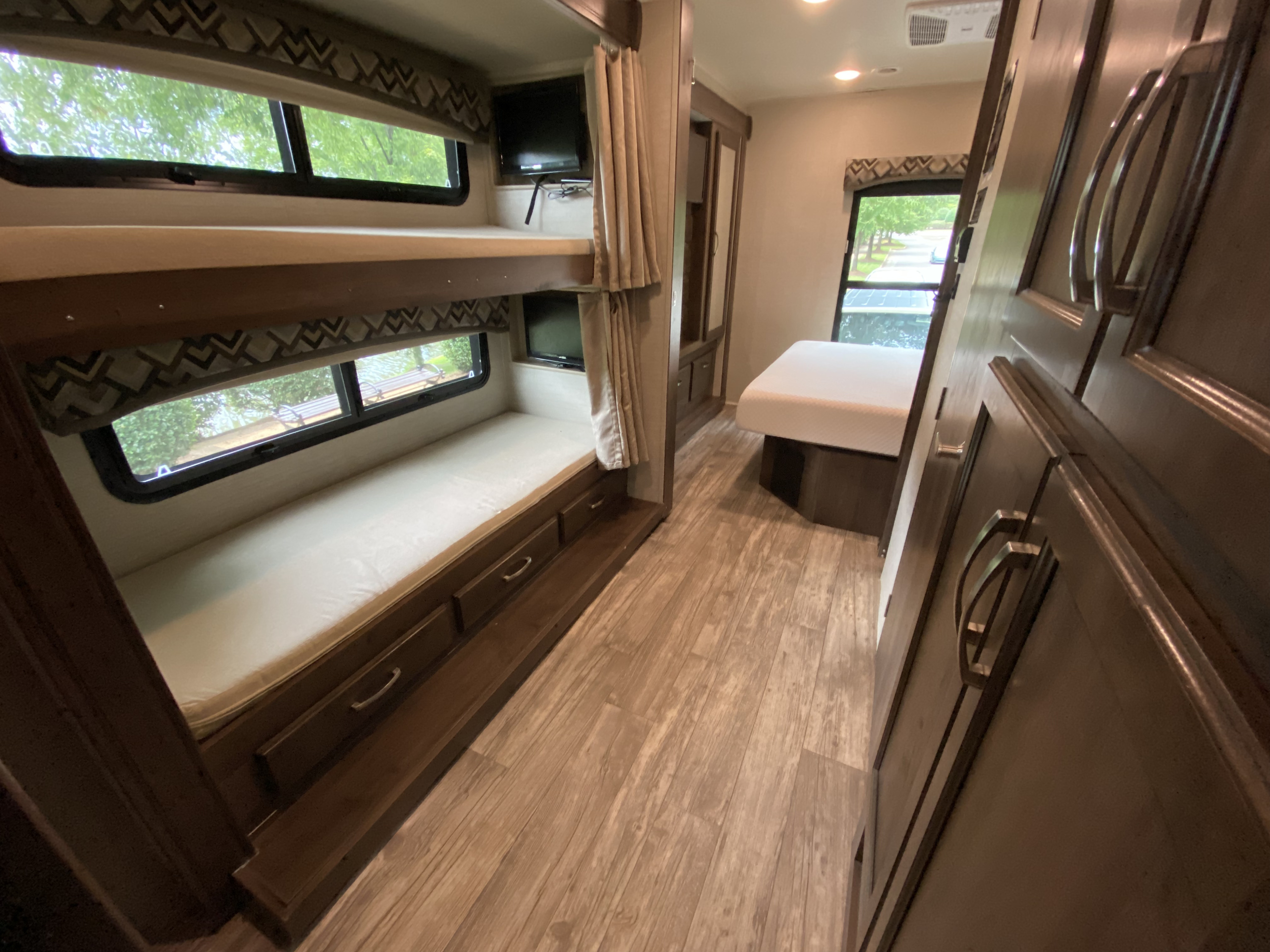 An RV rental interior featuring comfortable bunk beds, a main bed, wood floors, and bright windows with green views.. Jayco Jay Series 2018